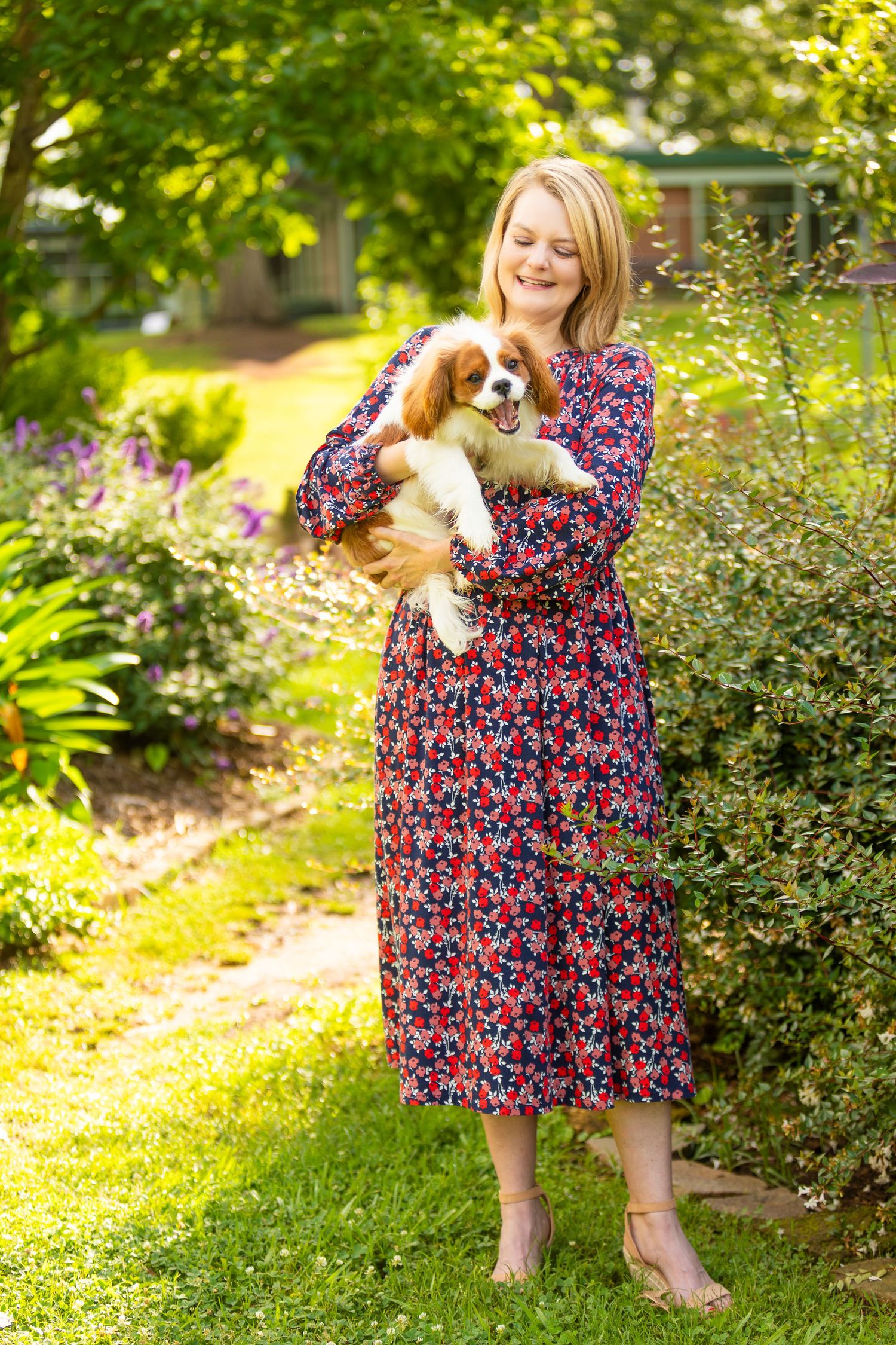 A woman in a floral dress plays outdoors in the garden with a playful King Charles Cavalier Spaniel puppy.