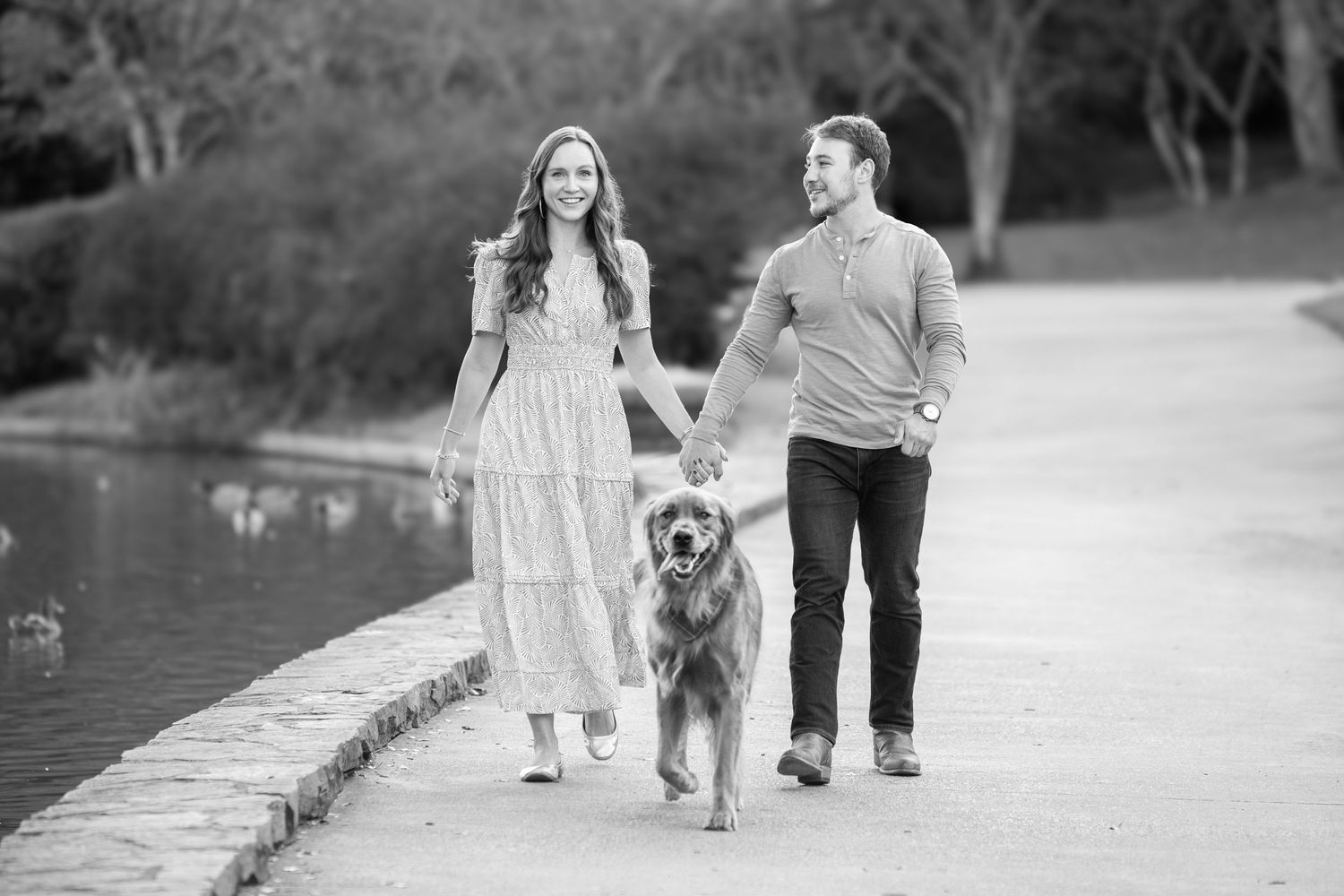 A couple walks their golden retriever dog along a lakeside path in a black and white photograph.