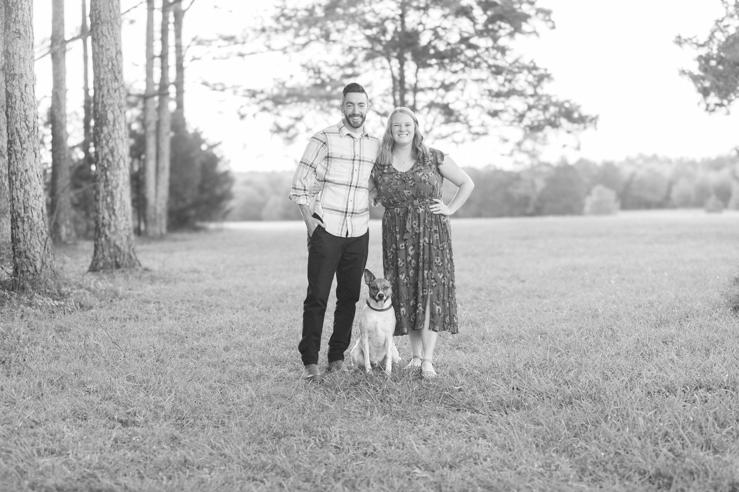 Black and white portrait of couple with their dog in tree-lined field.