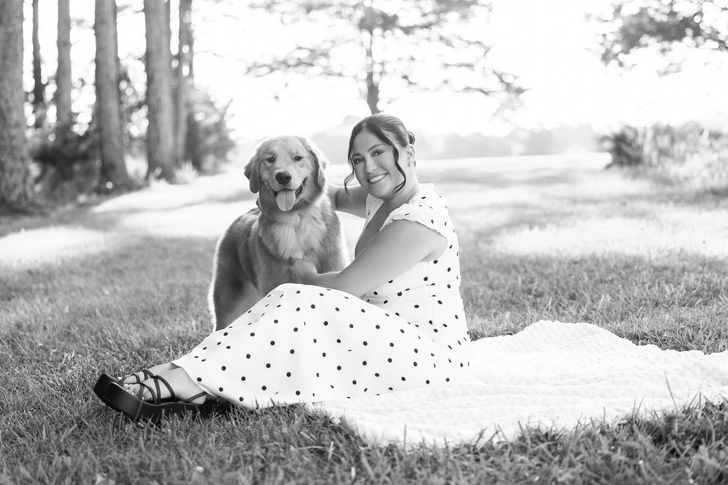Woman in polka dot dress sits smiling with golden retriever on grass near trees in black and white photo.