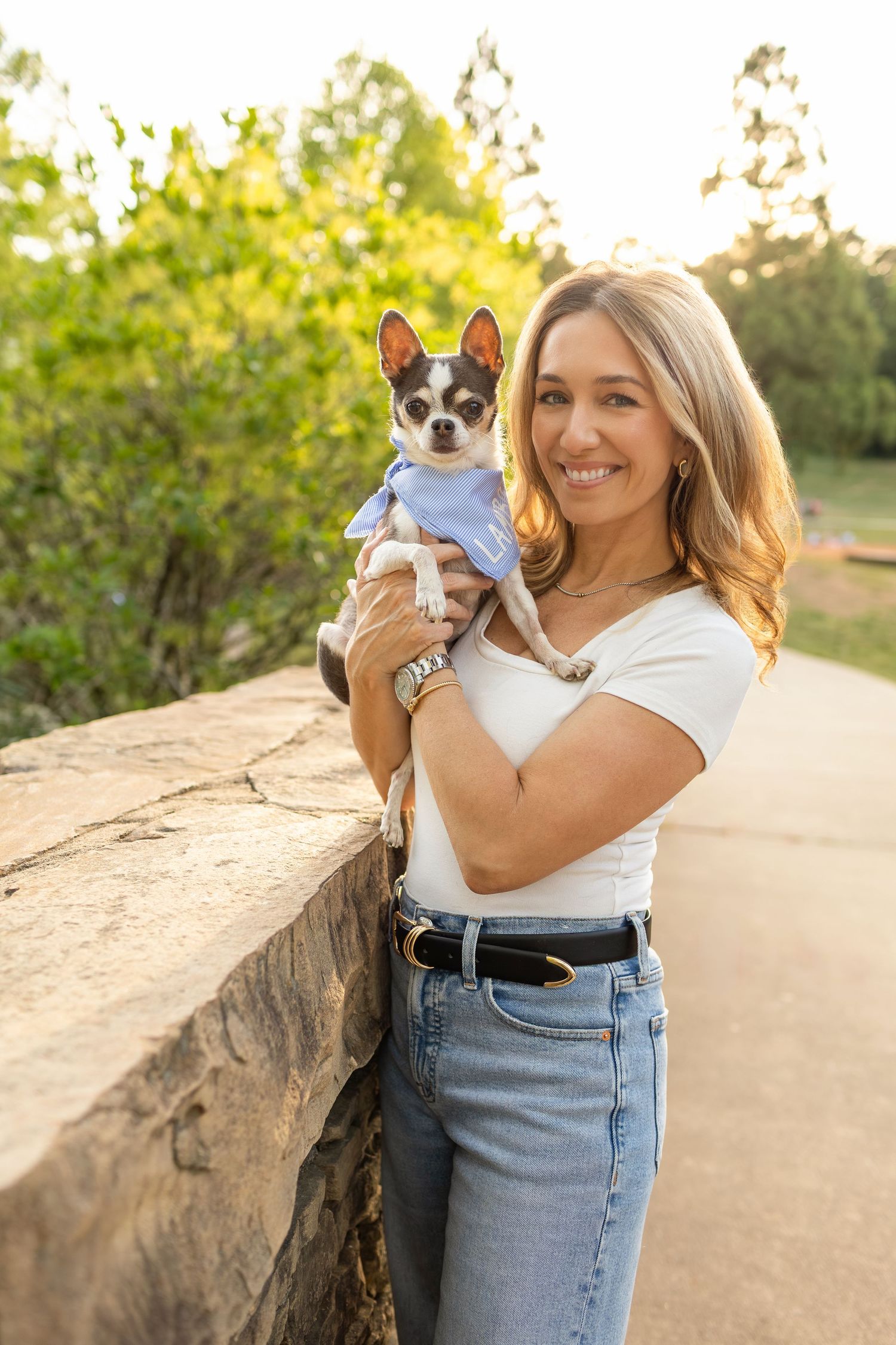 Smiling woman holding a small Chihuahua dog in a blue shirt outdoors in a sunny park setting.