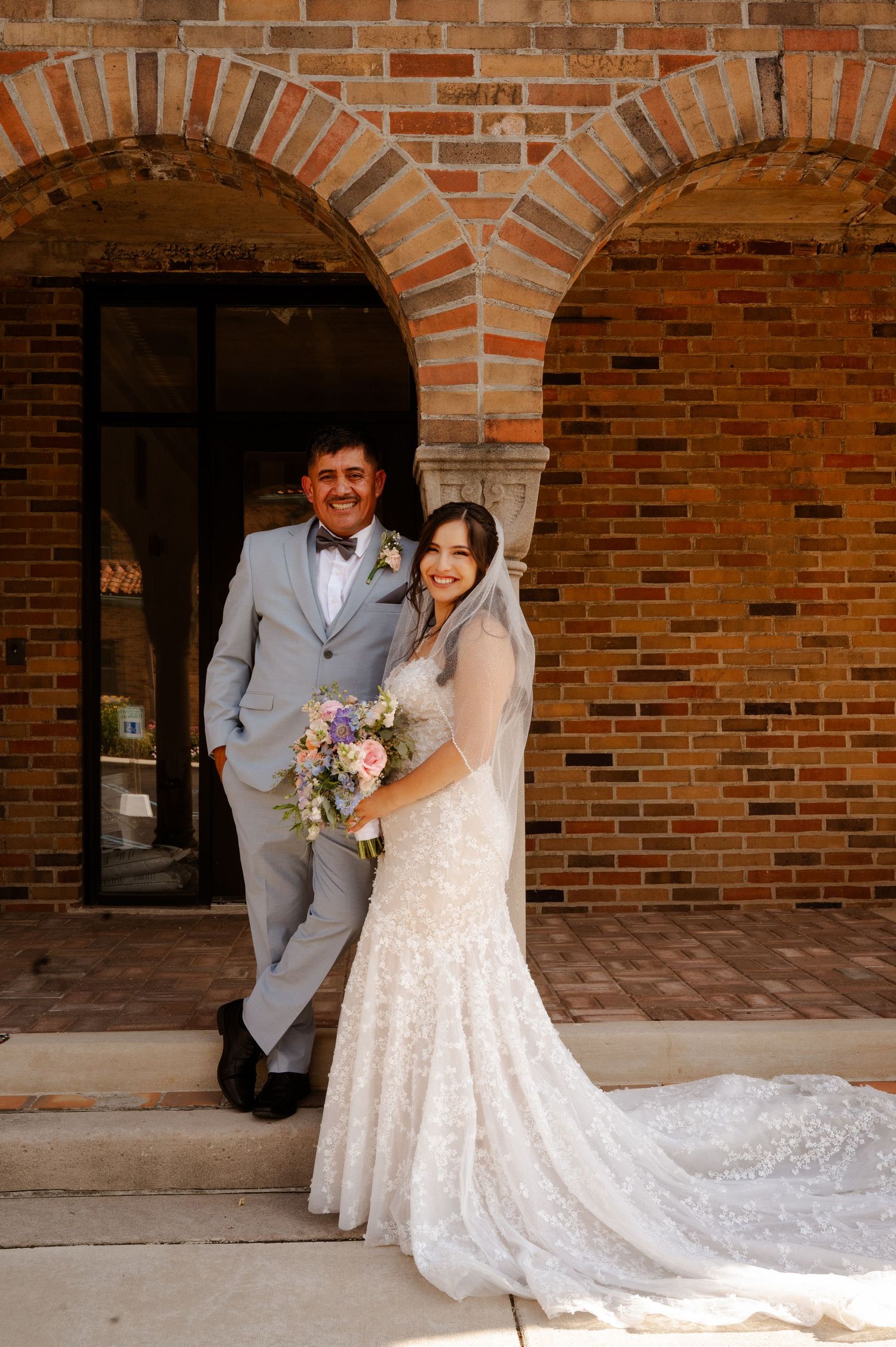 Bride in lace gown holding bouquet stands with groom in gray suit before brick arched doorway at wedding venue.