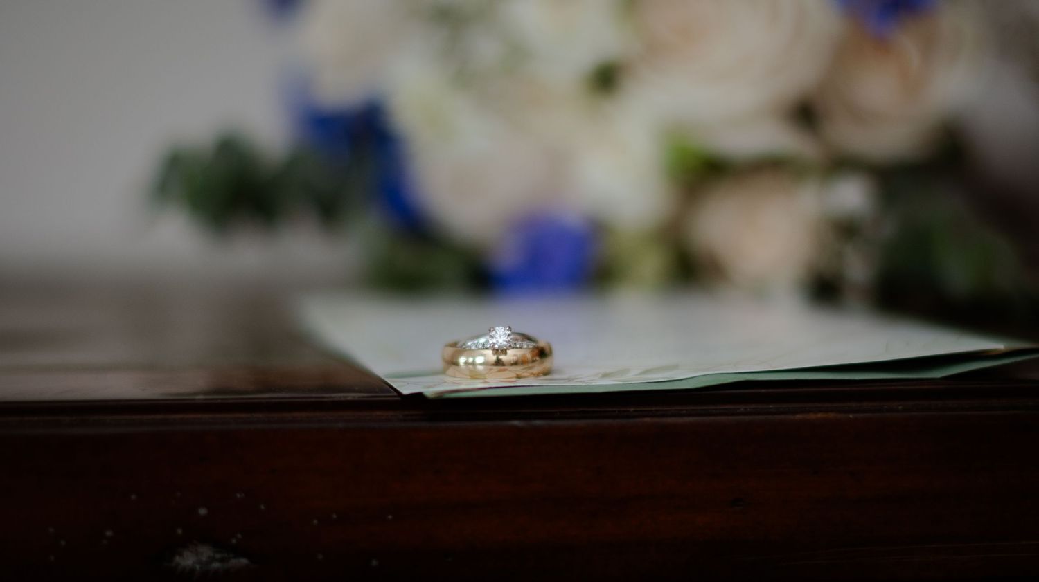 Diamond engagement ring and wedding band rest on top of a turquoise surface with soft floral decorations in background.