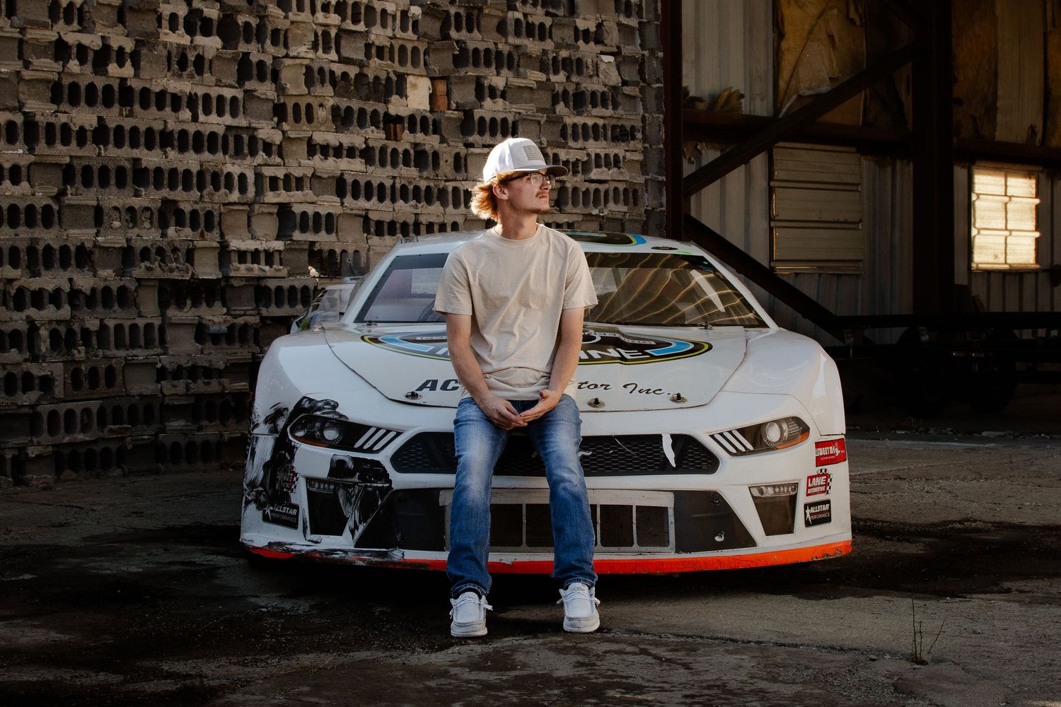A person sits on the hood of a white customized sports car with racing decals parked against a brick wall.