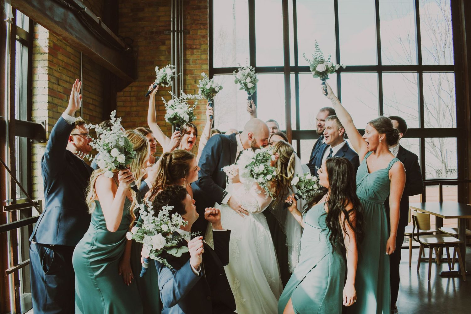 Wedding party celebrates with arms raised in industrial venue with tall windows.