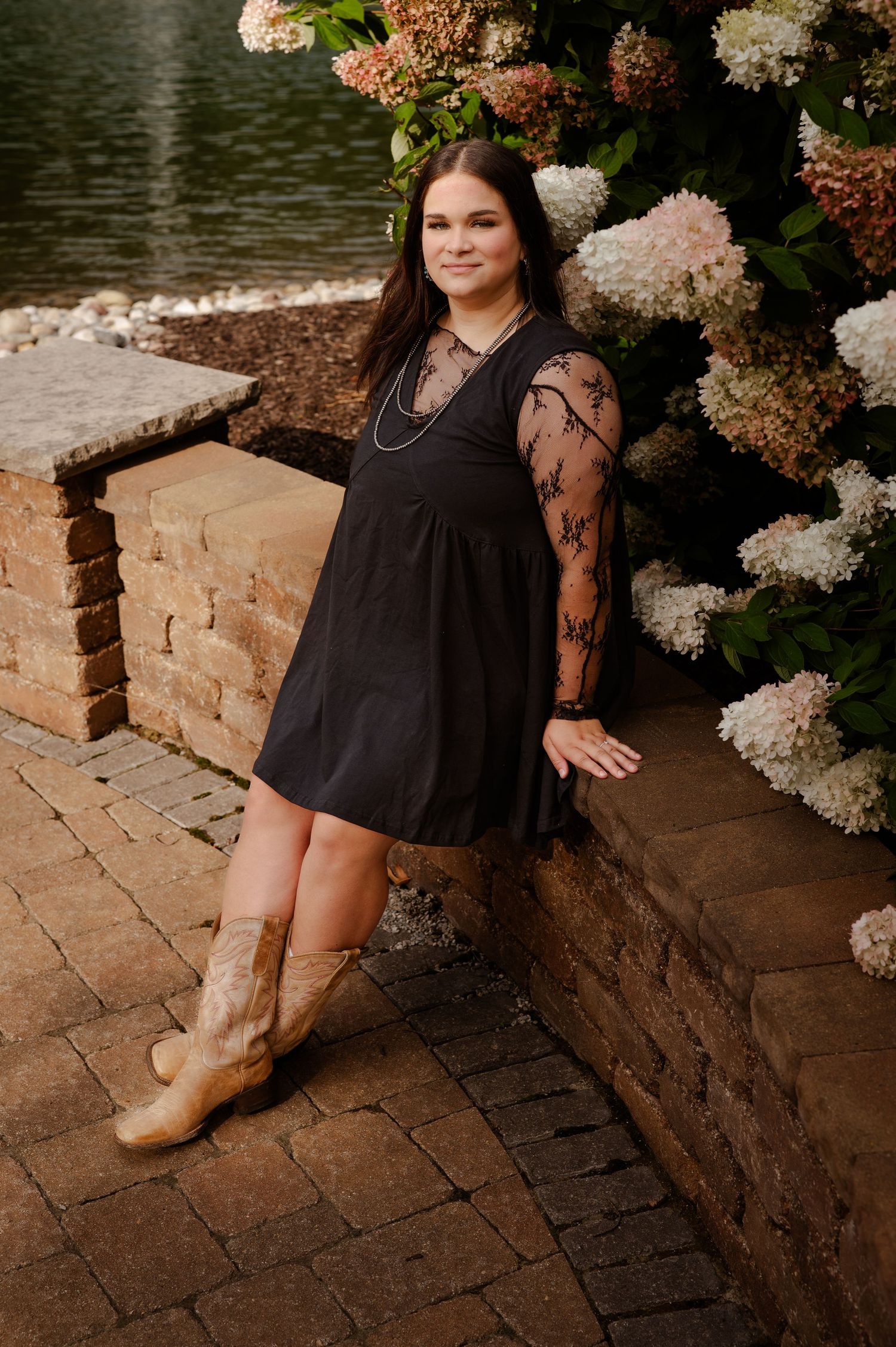Autumn outdoor portrait by stone wall with hydrangeas and wearing a black dress with lace sleeves and tan boots.