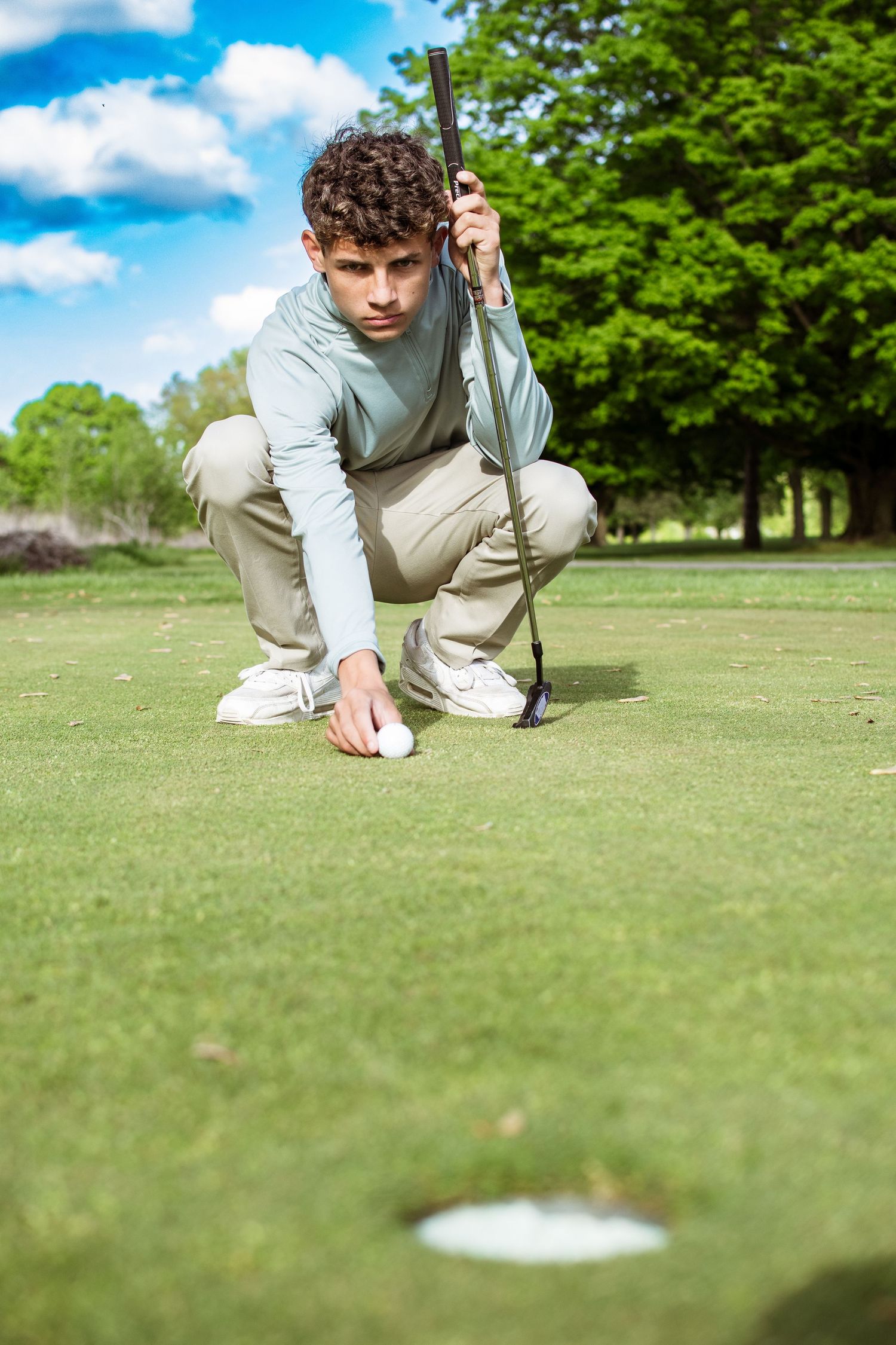 Golfer crouches on putting green to line up shot while holding putter on sunny day at golf course.