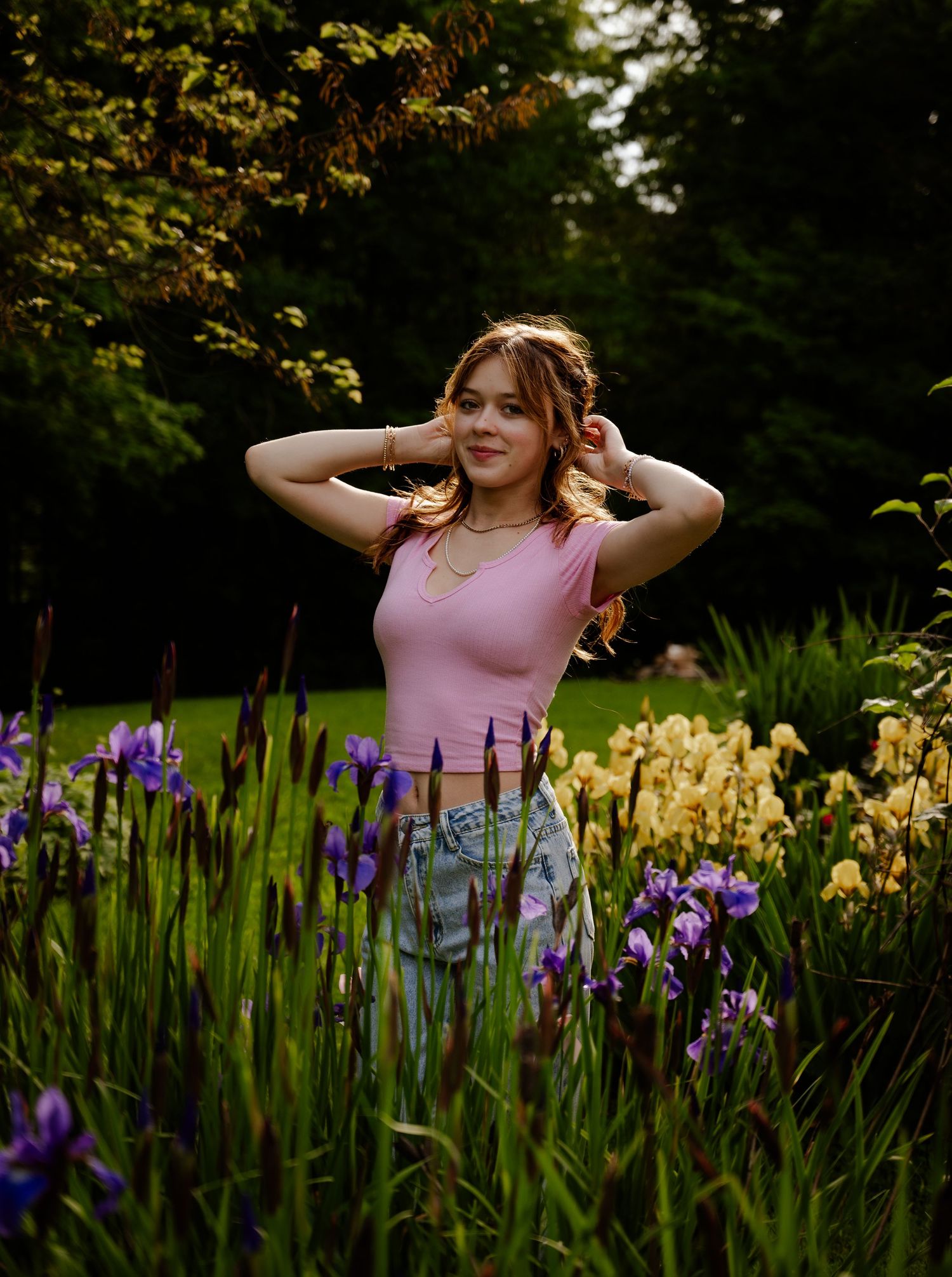 Someone in a pink tank top poses in a garden with blooming purple and yellow iris flowers on a summer evening.