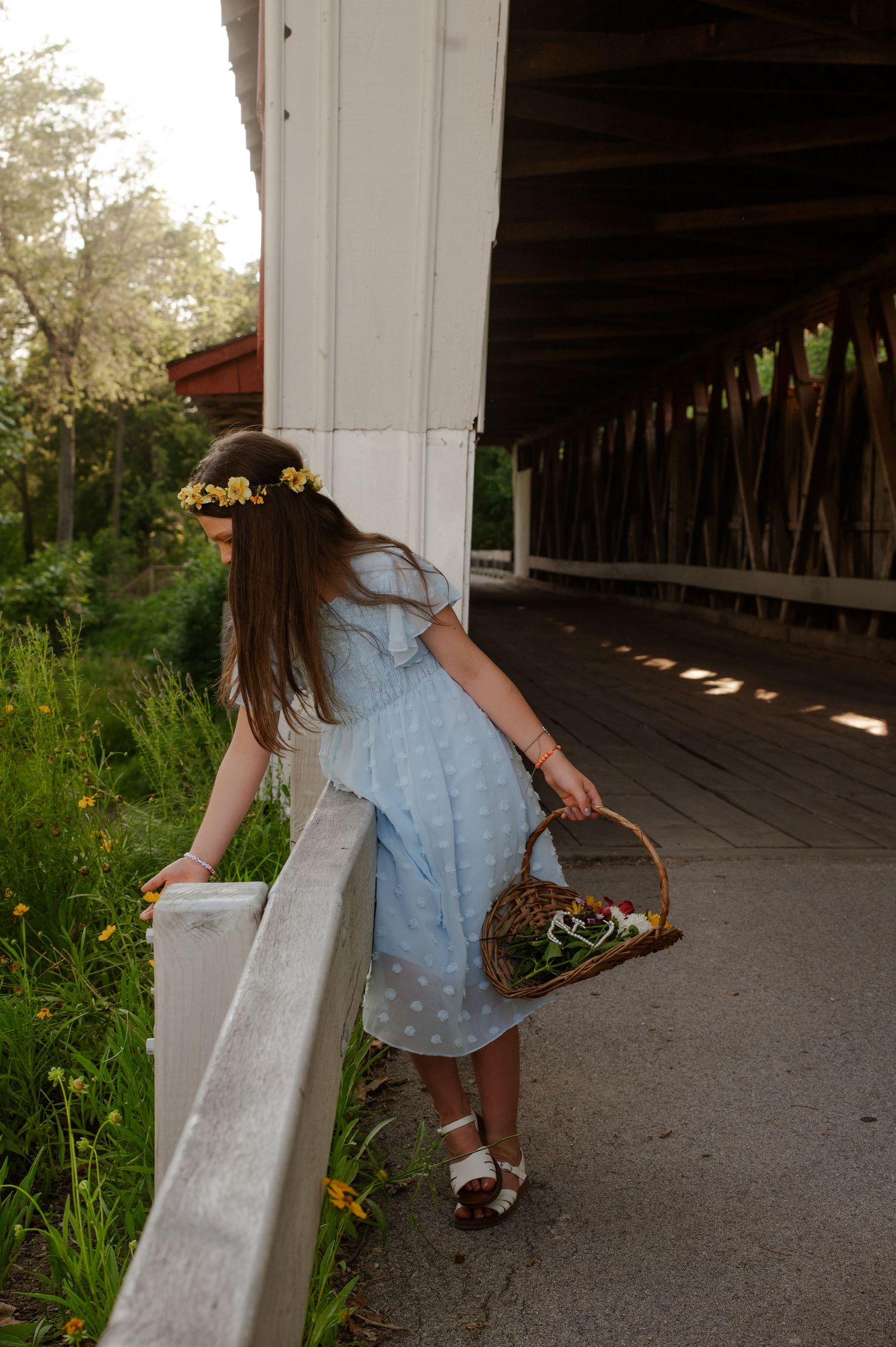 Someone in a blue dress and floral crown holds a basket while standing at the edge of a covered wooden bridge.