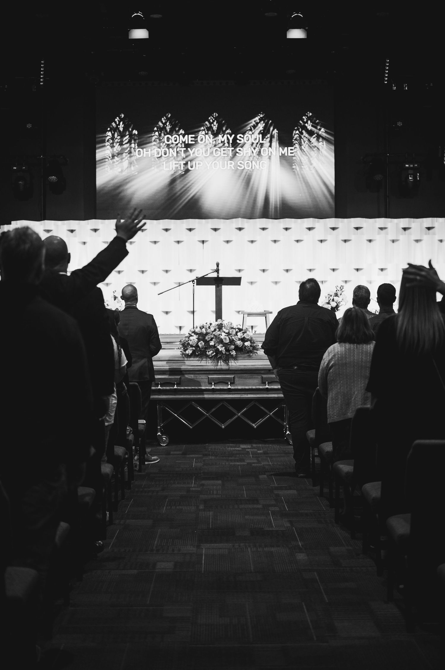 Black and white photo of a church service with raised hands in silhouette and a cross-adorned casket in front.
