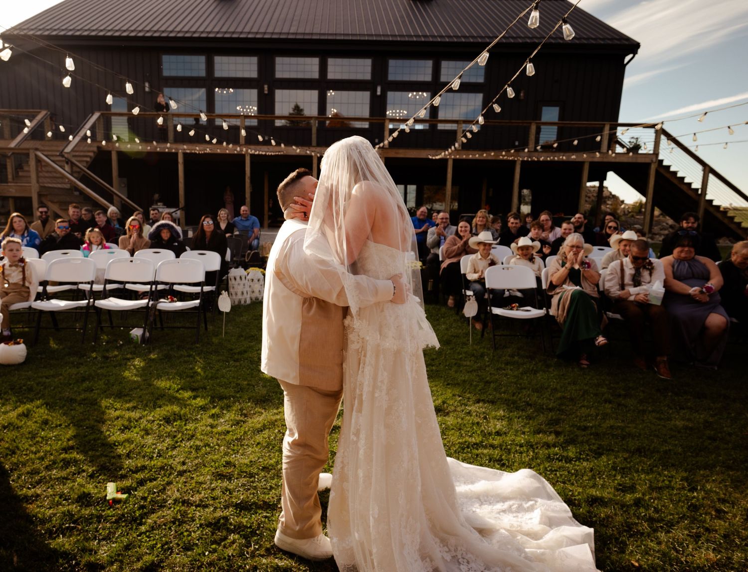A romantic outdoor wedding moment with guests seated on white chairs at a rustic venue.