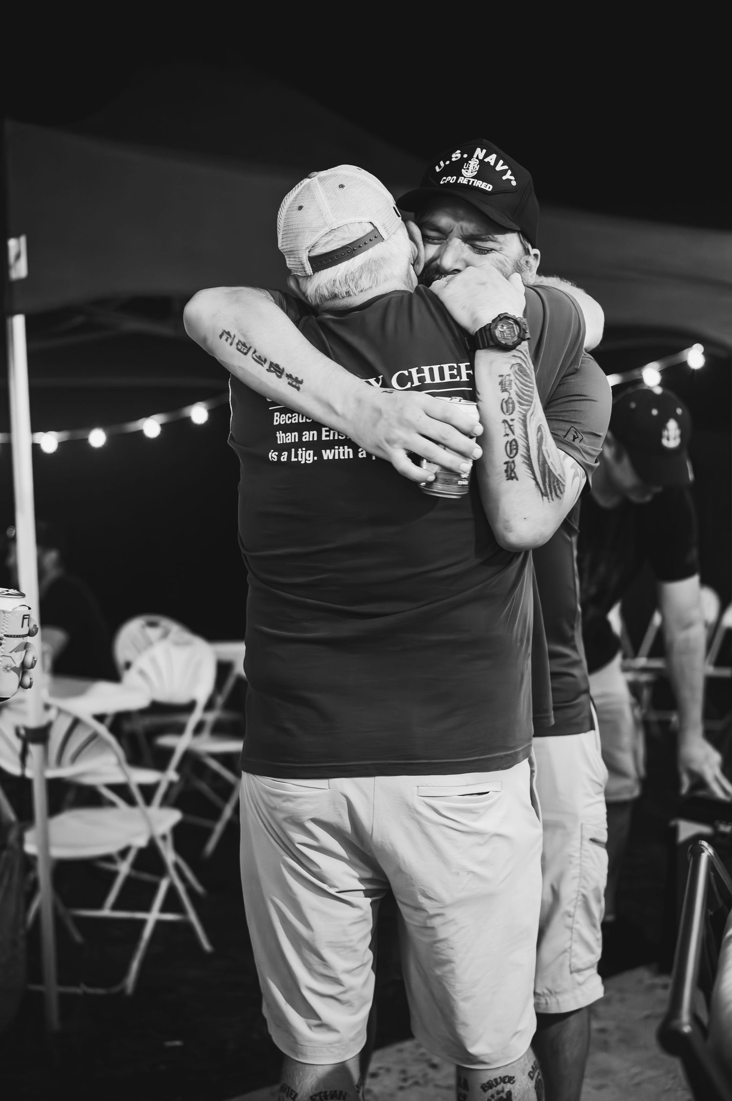 Two people sharing emotional embrace at outdoor event with folding chairs visible.