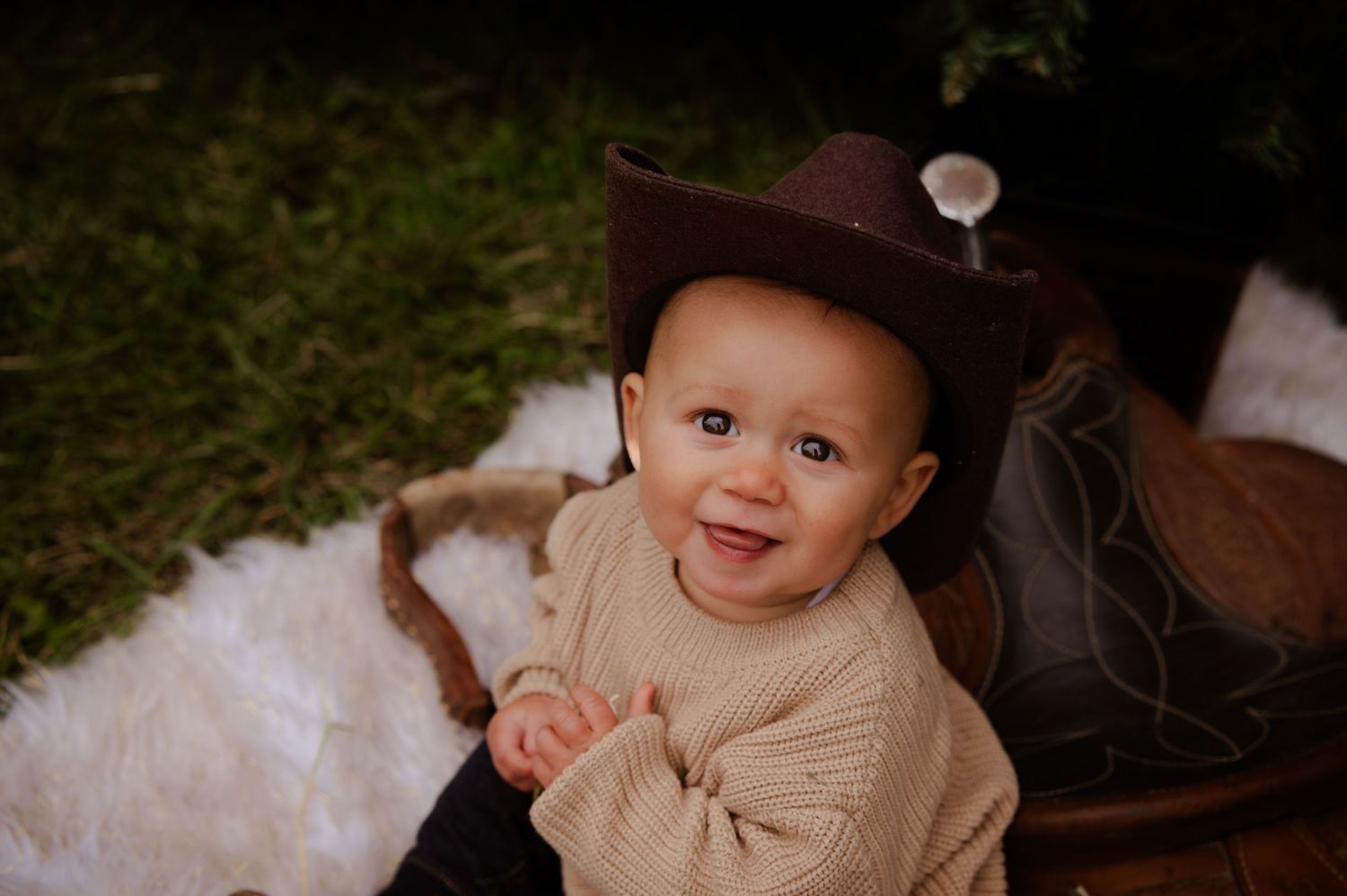 Baby wearing cowboy hat smiles while sitting on fluffy blanket outdoors.