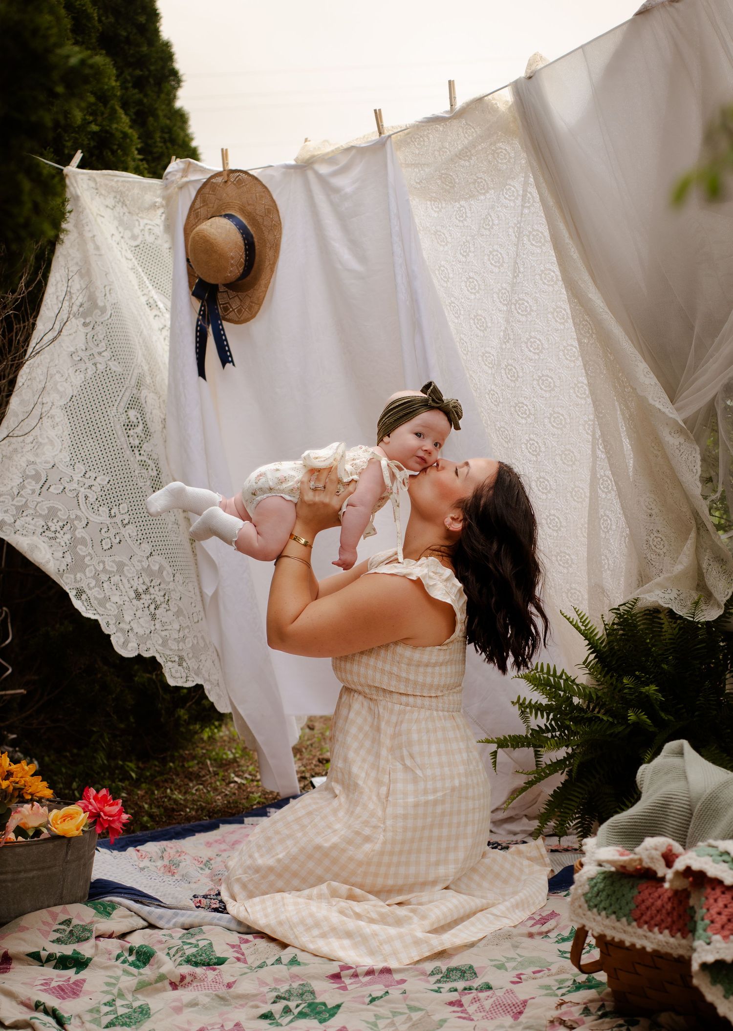 Mother and baby laying on picnic blanket with white laundry drying on clothesline in outdoor garden setting.