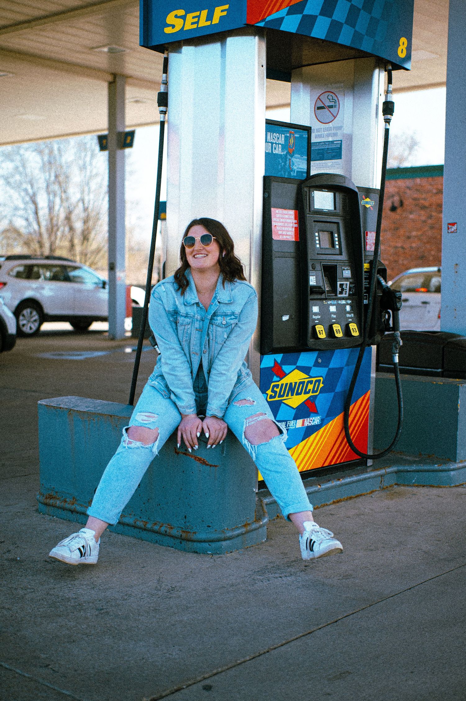 Someone in ripped jeans and denim jacket sits casually on gas pump base at self-service station on sunny day.