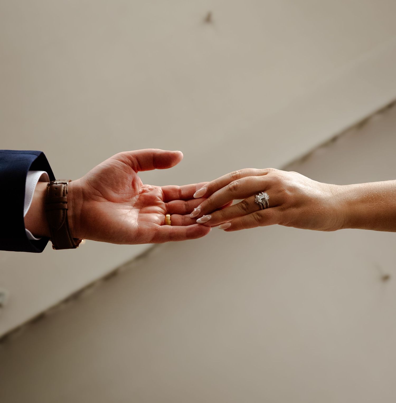 Two hands reaching toward each other, one wearing a suit and bracelet, the other adorned with a diamond engagement ring.