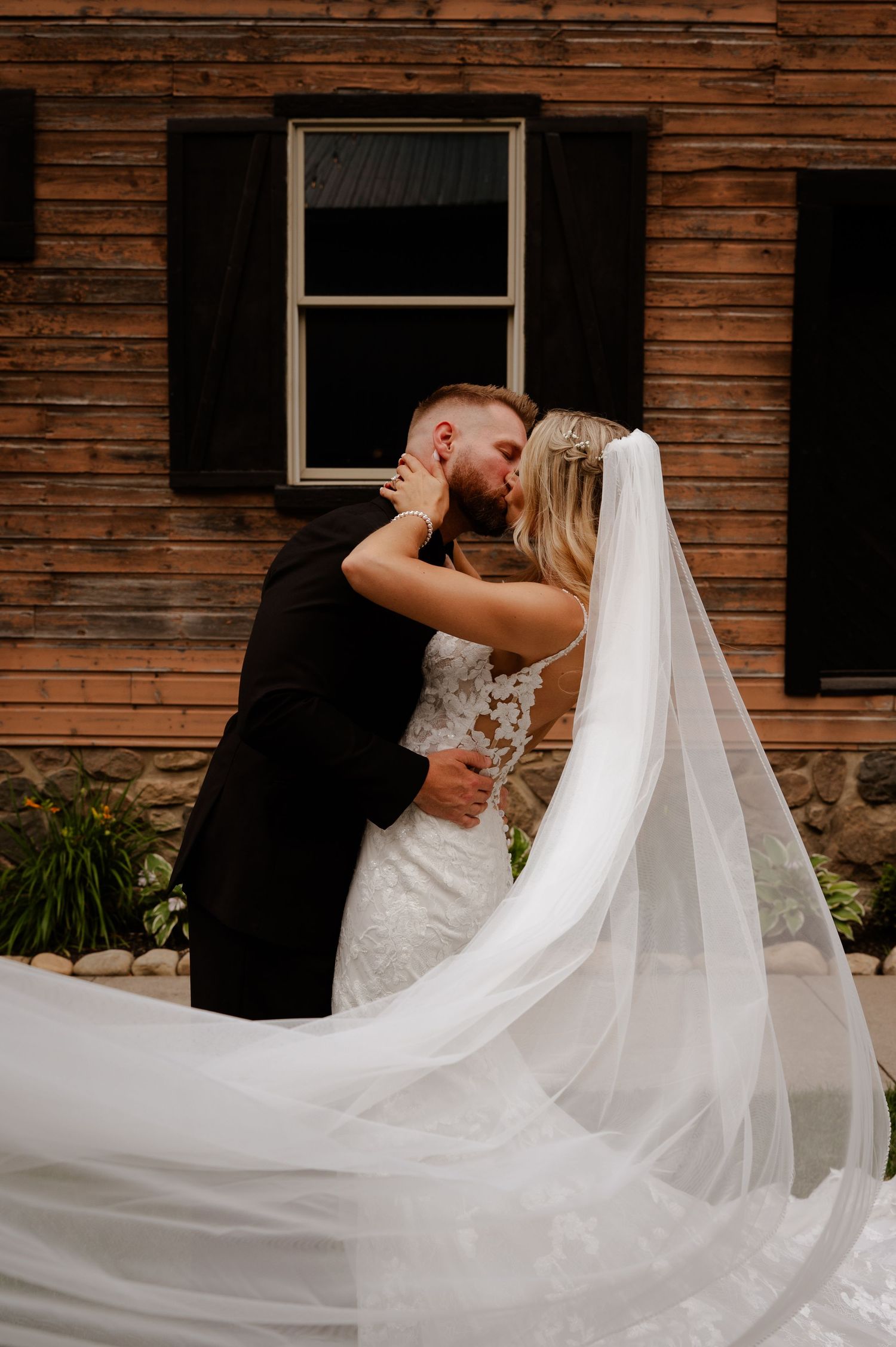 Bride and groom share a romantic kiss outdoors, her cathedral veil flowing, against a rustic wooden barn backdrop.