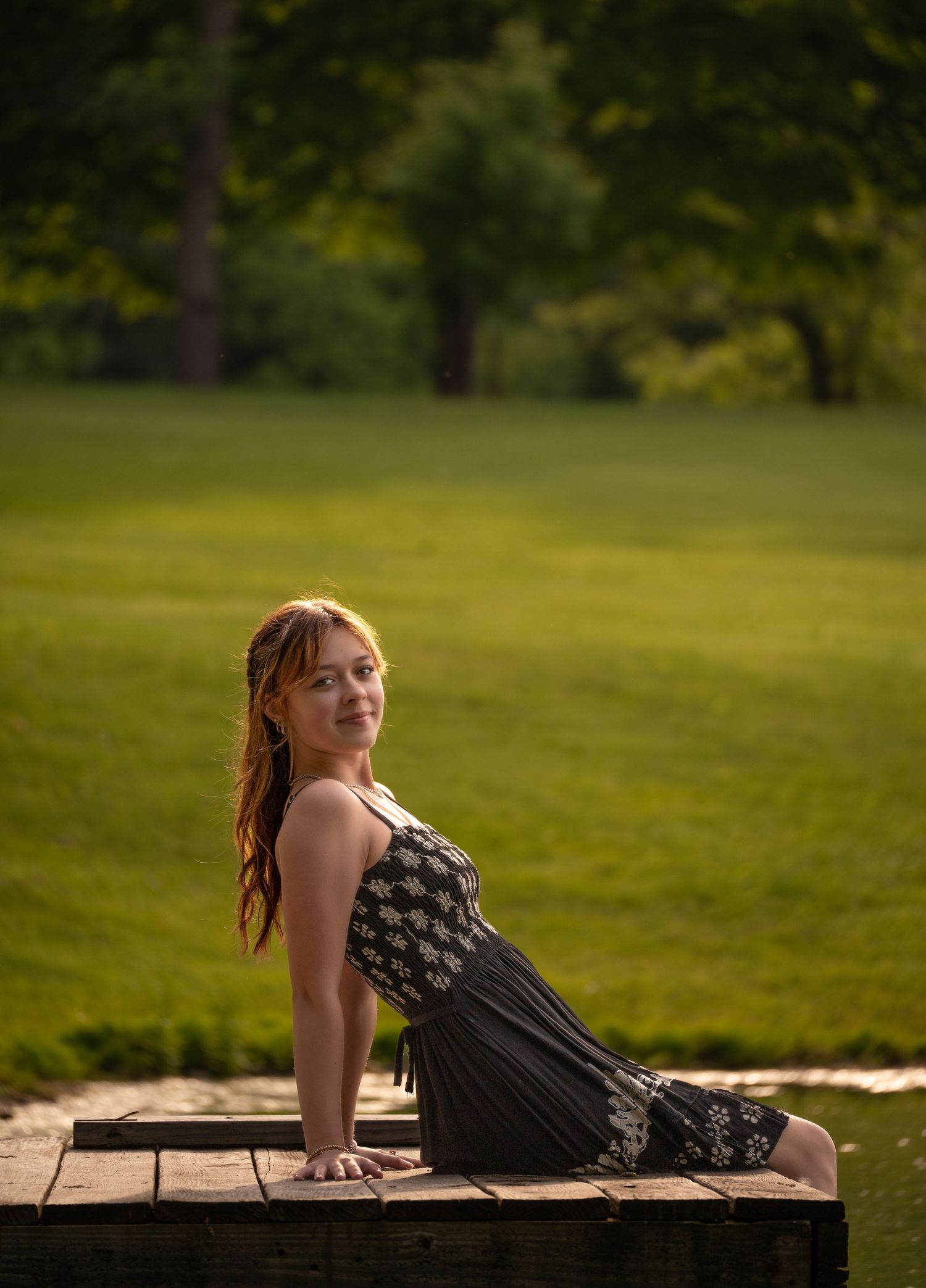 Person in a black patterned dress poses on wooden deck with green lawn and trees in soft evening light.