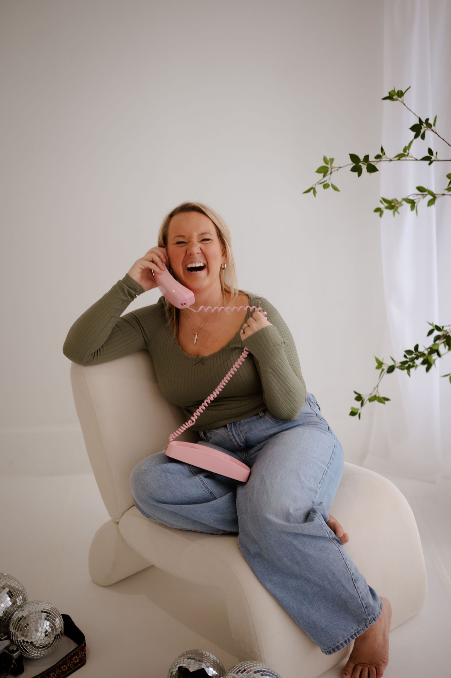 Laughing blonde woman in olive top and jeans sits in white chair holding pink retro phone with pink crossbody bag.