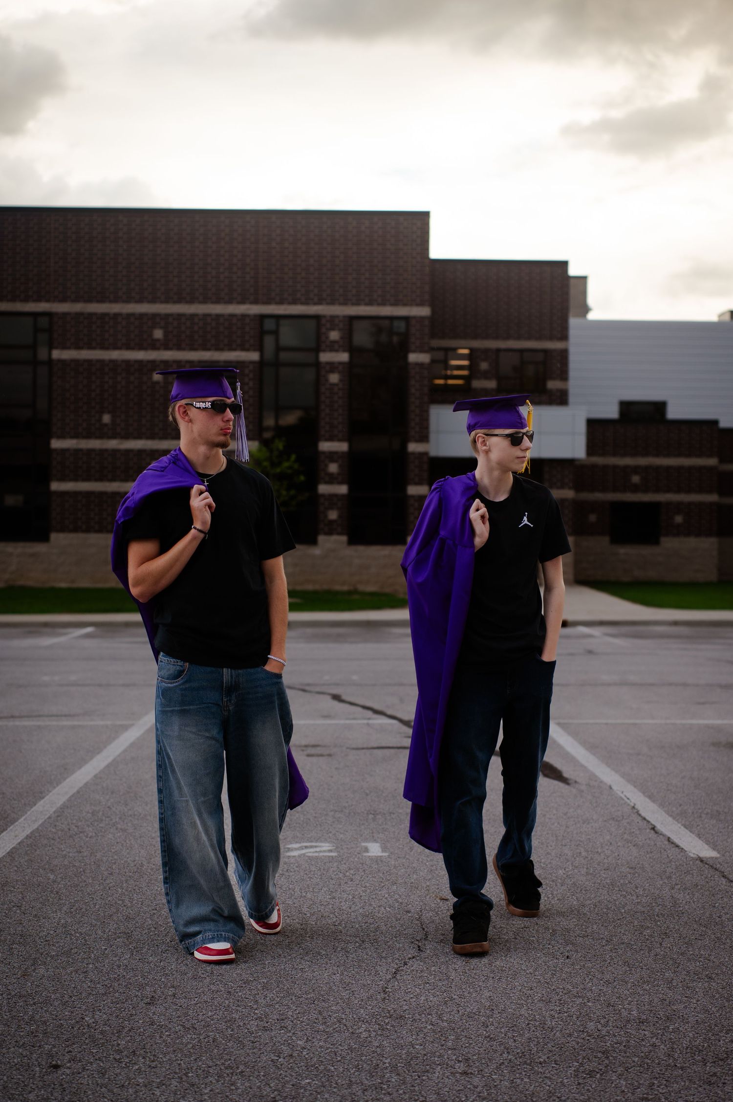 Two people walking through a parking lot wearing purple graduation gowns and black clothing.