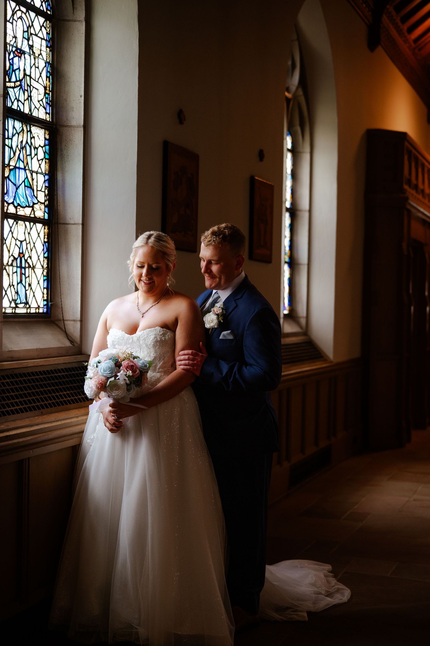 Bride and groom share a romantic moment in a church with stained glass windows, bride holding a floral bouquet.