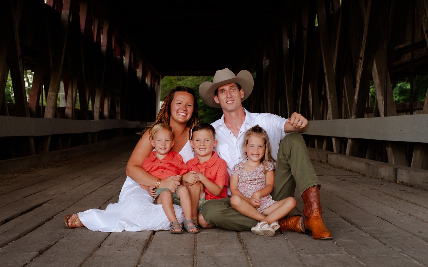A family sits together on a wooden covered bridge floor wearing coordinated coral and earth-toned outfits.