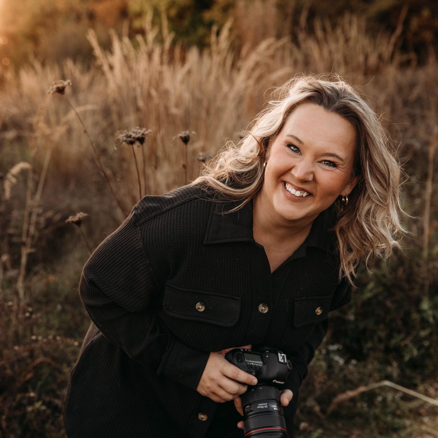 A photographer with a DSLR camera laughs joyfully while taking photos in a golden fall field with wheat stalks.
