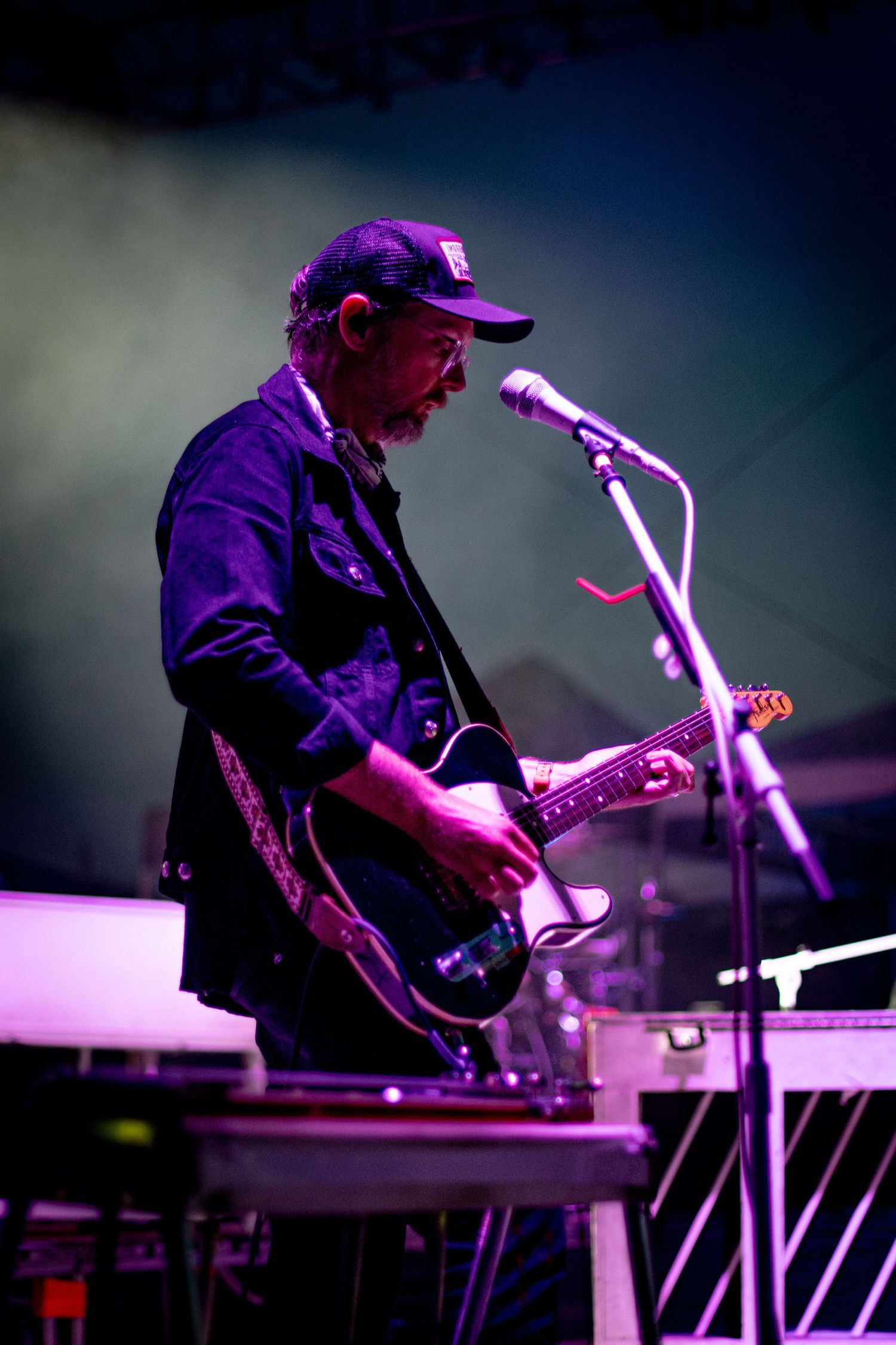 A musician performs on stage with a guitar and microphone under dramatic purple lighting during a concert performance.