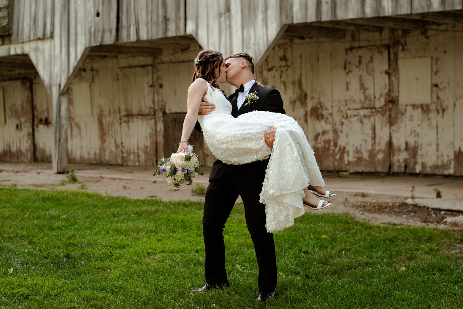 A groom lifts his bride in a romantic pose against a rustic barn backdrop.