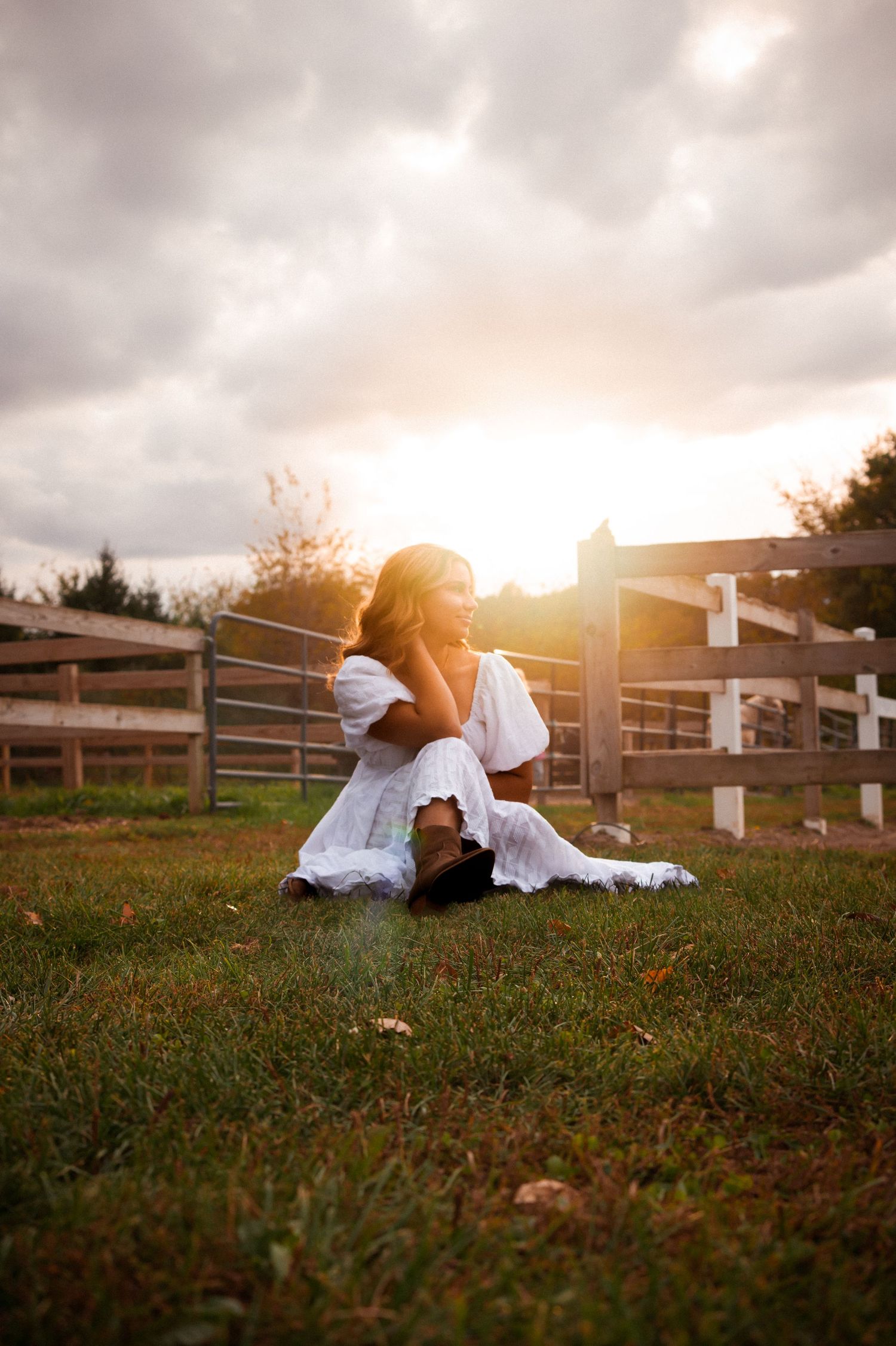 Ethereal portrait in white dress sitting on grass during golden hour with fence in background.
