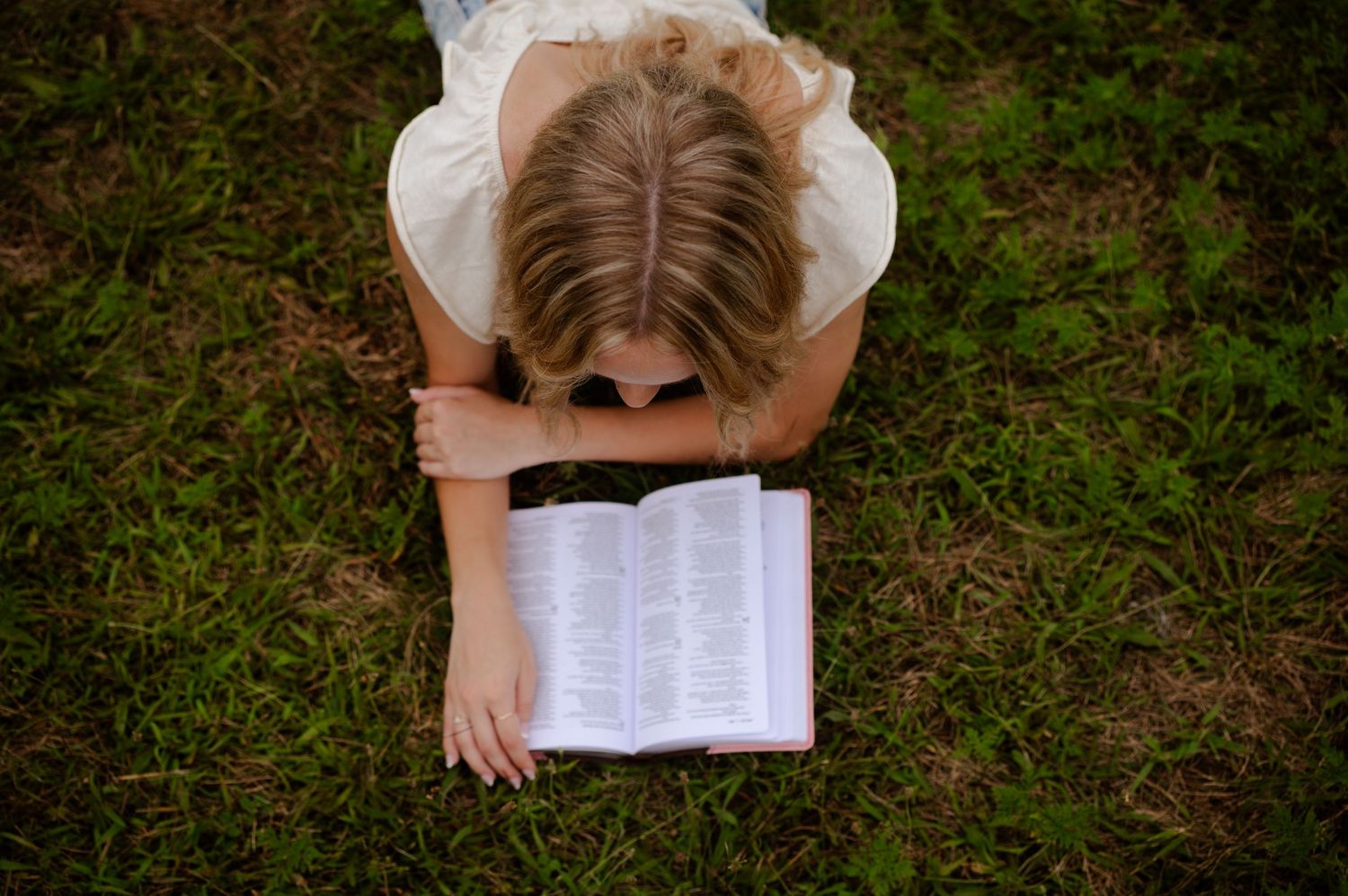 A person lies in the grass while reading from a book held overhead against a natural green background.