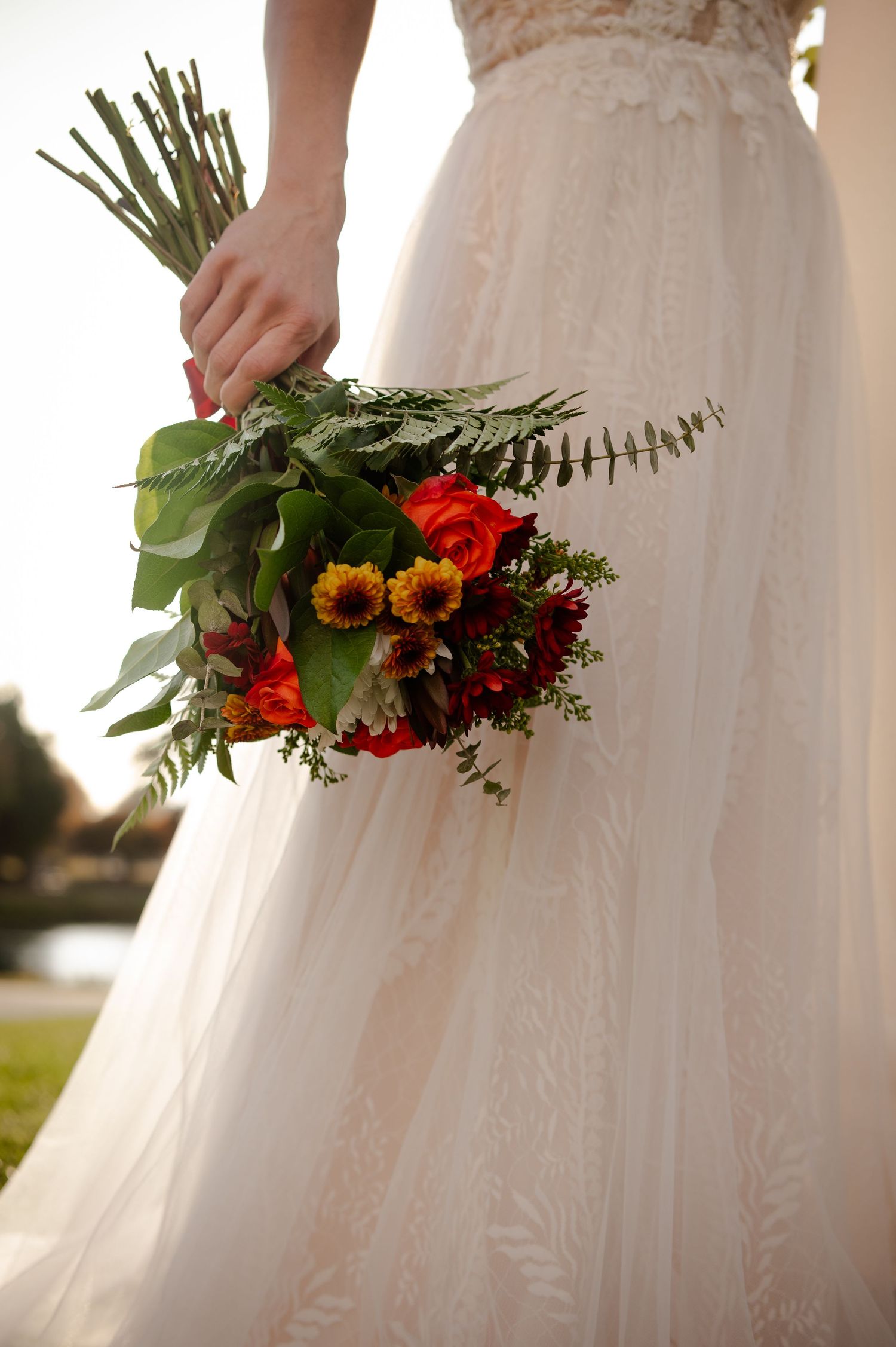 Bride holding vibrant fall wedding bouquet with sunflowers and red flowers against white dress.