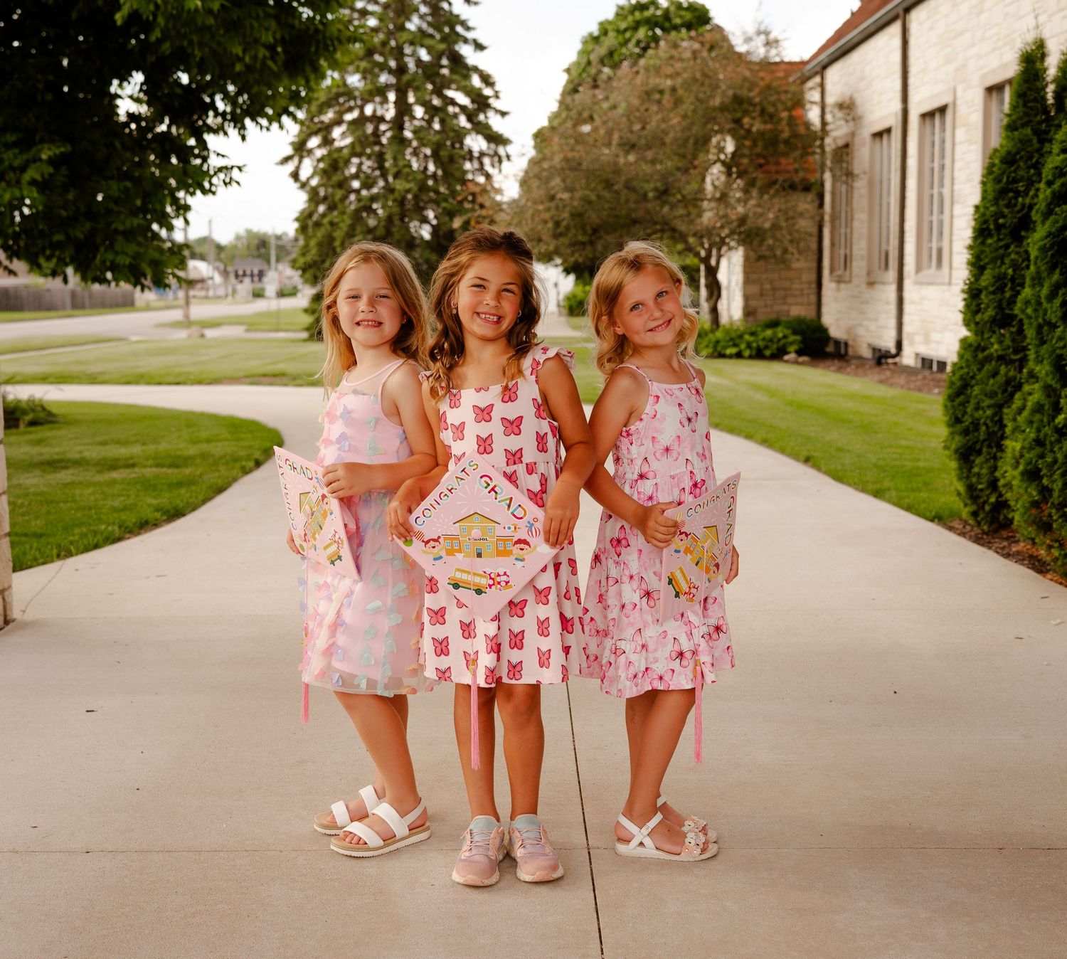 Three young girls in matching pink dresses pose together on a sidewalk, smiling and holding bags and a book.