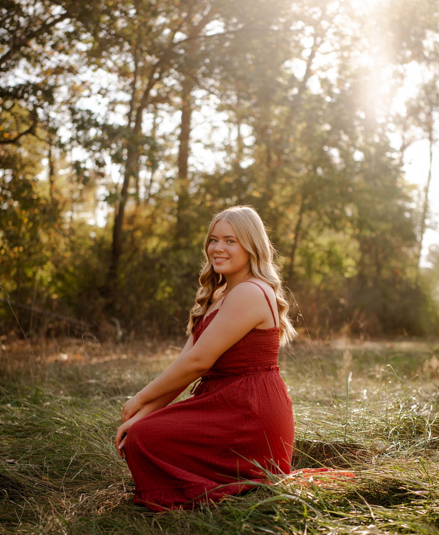 A person in a flowing red dress sits gracefully in a sunlit meadow during golden hour.