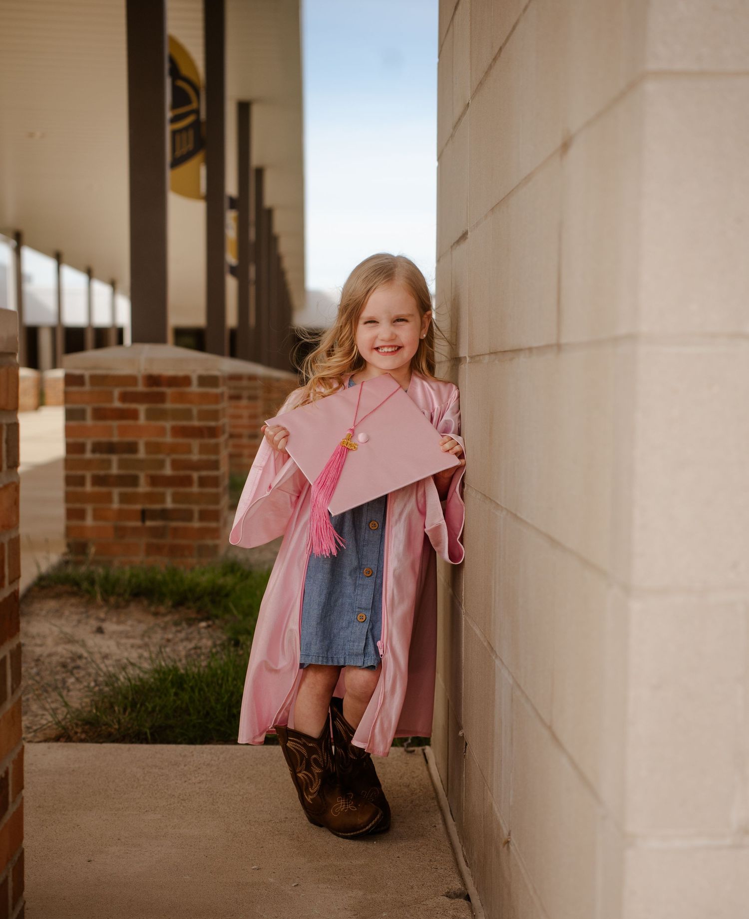 A child in a pink coat and denim dress stands against a wall in a sunny outdoor corridor.