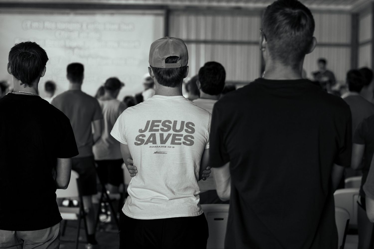 A person wearing a white 'Jesus Saves' t-shirt stands among a crowd in a church or meeting hall.