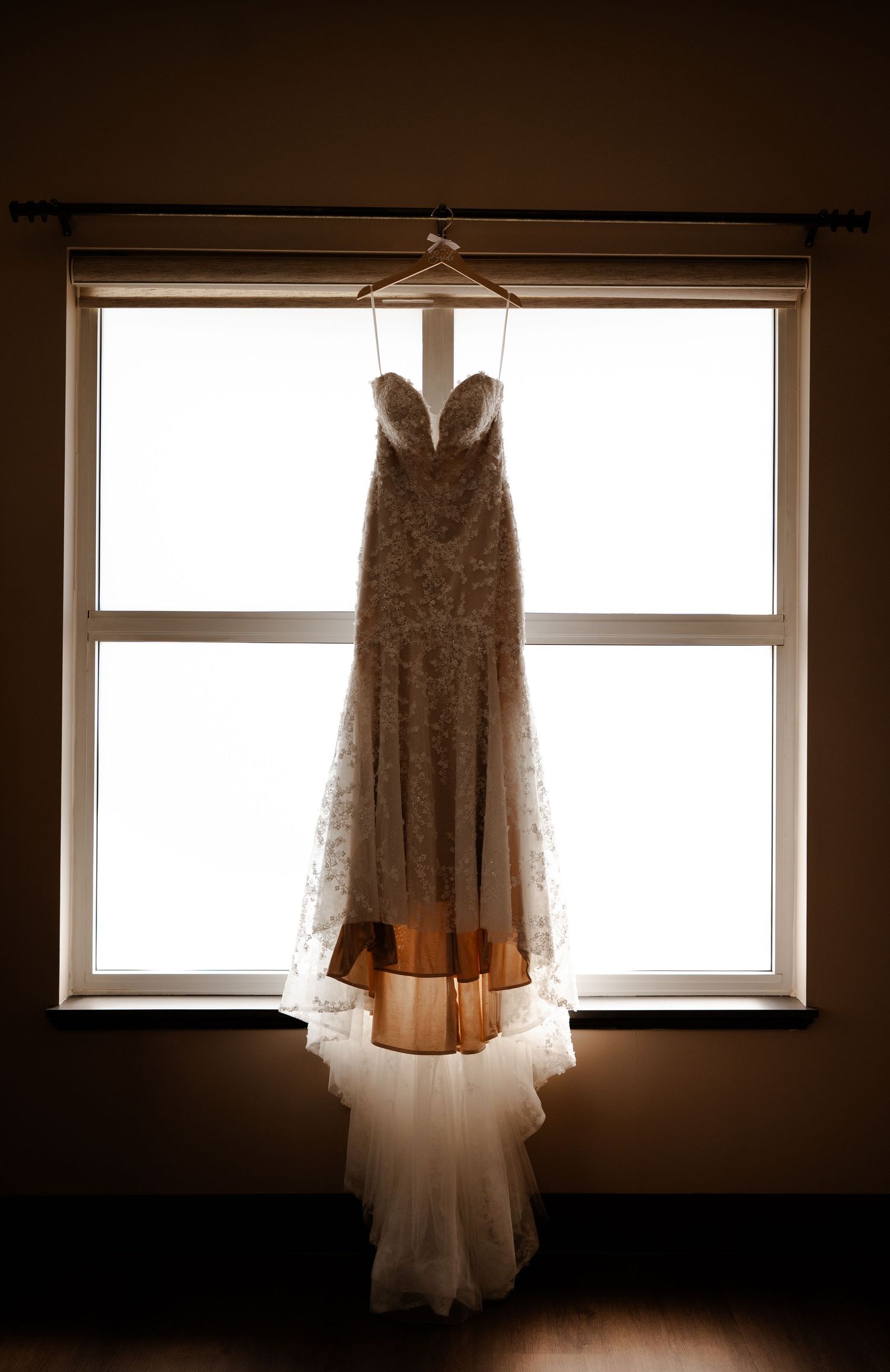 Elegant lace wedding dress hanging on a wooden hanger in front of a bright window, showcasing its sweetheart neckline and train.