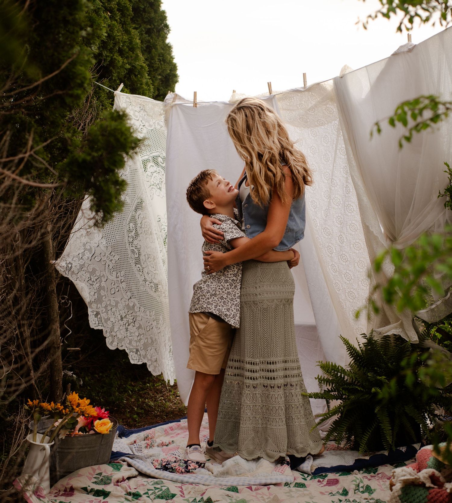 Two people embrace in a romantic moment against white linens hanging on a clothesline in an outdoor garden setting.