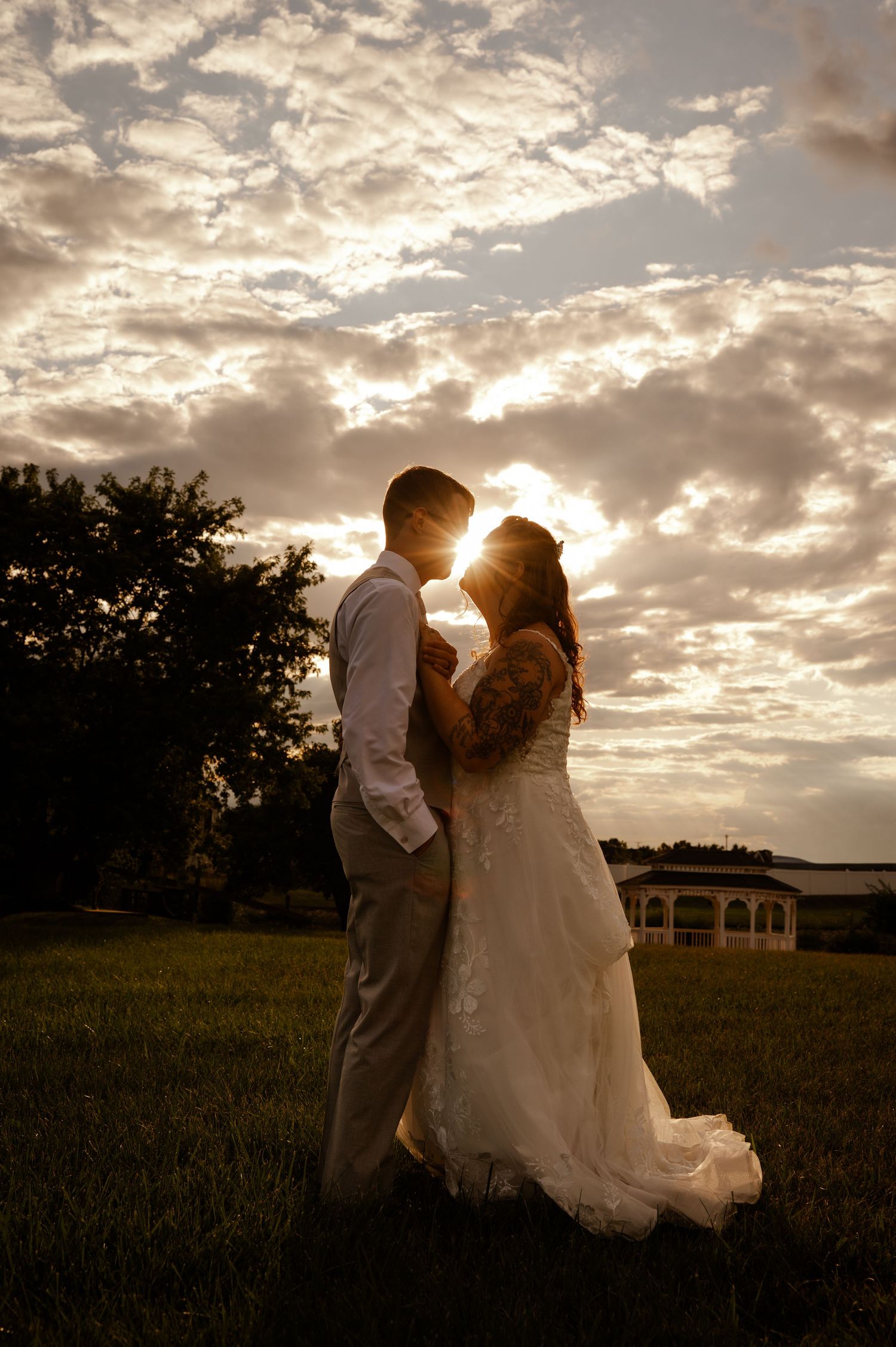 Silhouetted couple embraces in a romantic sunset field with dramatic cloudy sky and glowing sunlight in background.