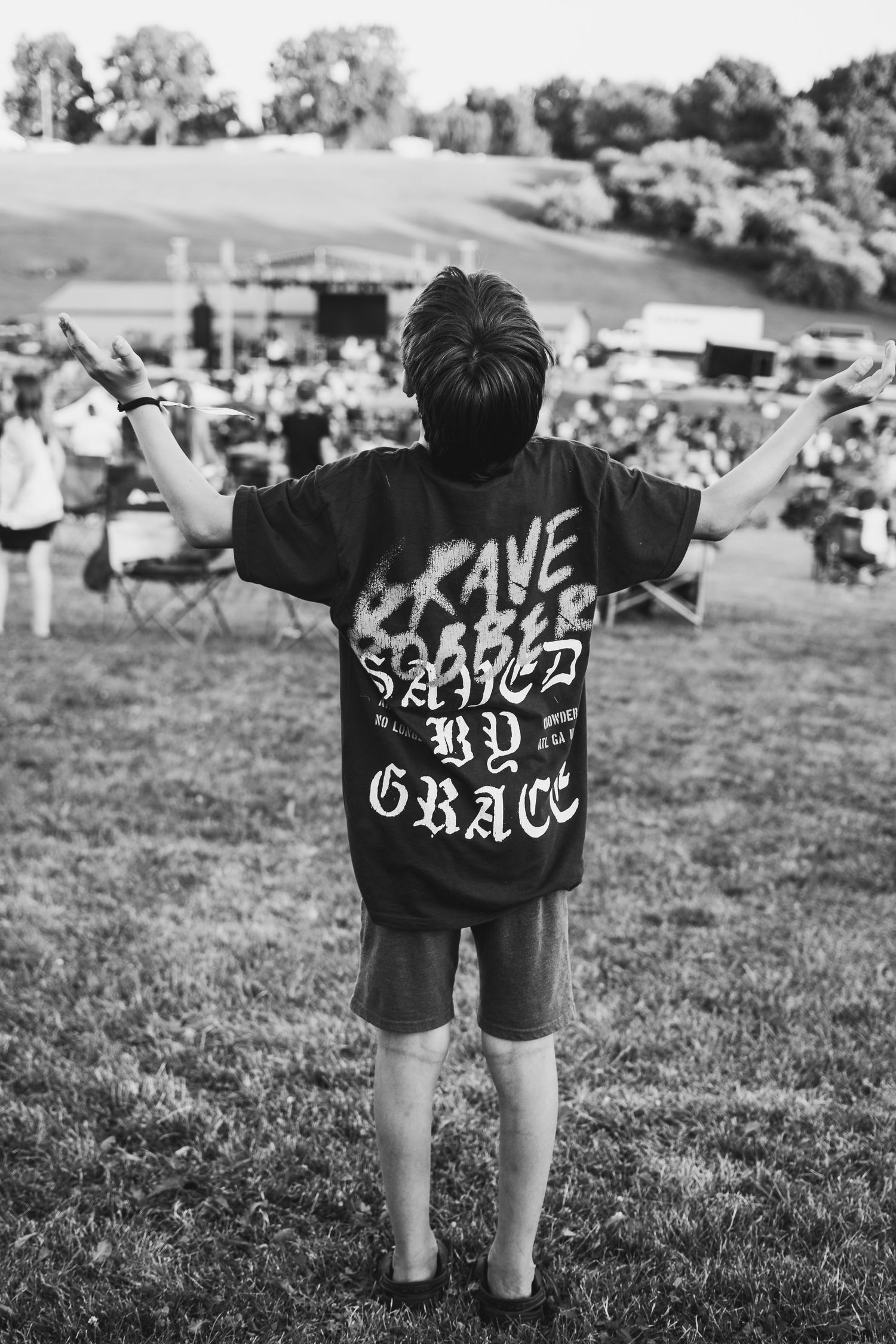 Black and white photo of someone in a 'Grave Robber Saved By Grace'' t-shirt at an outdoor festival on a grassy hill.