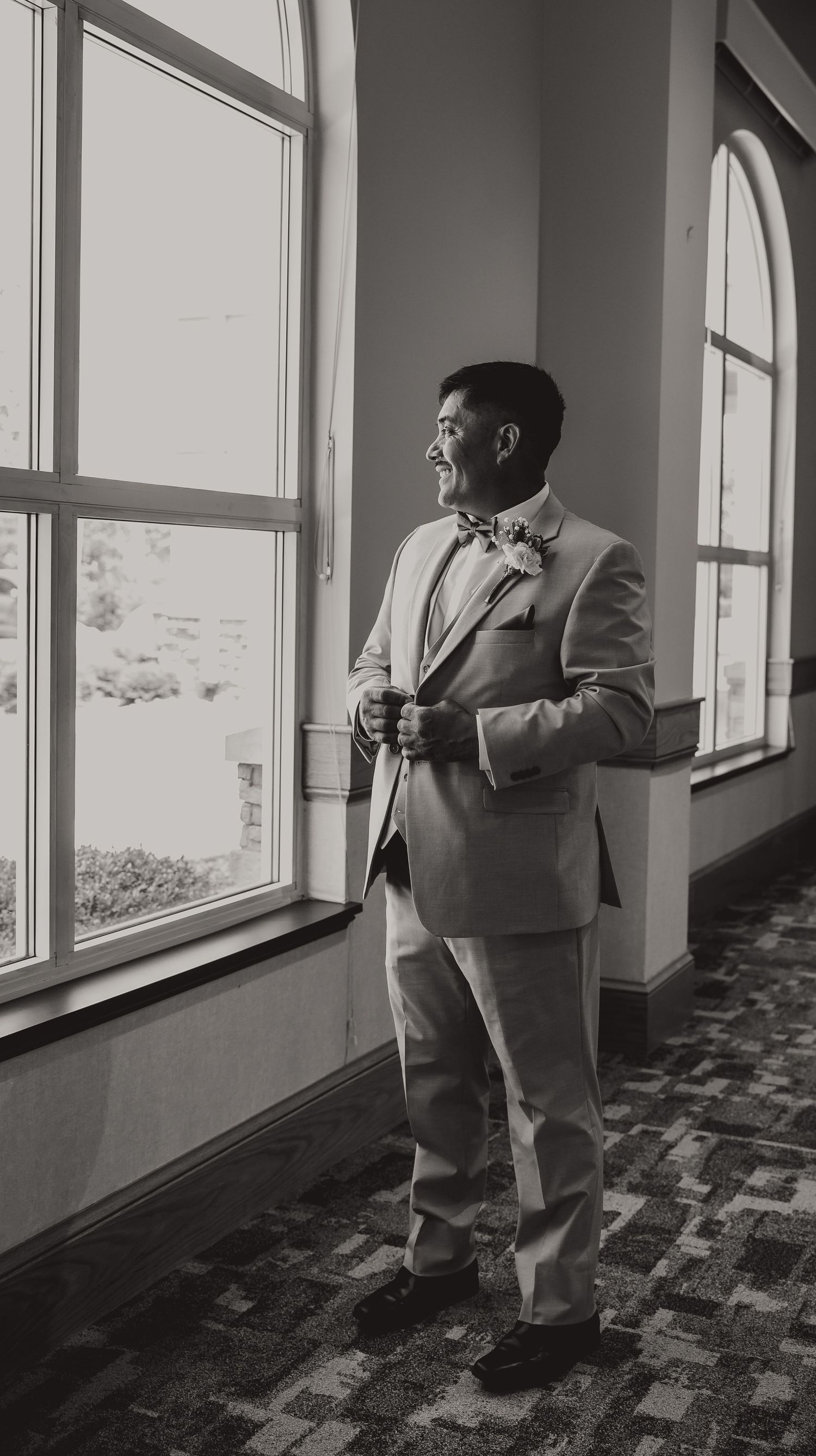 Black and white photo of a well-dressed man in a suit and bow tie standing by large windows in an elegant venue.