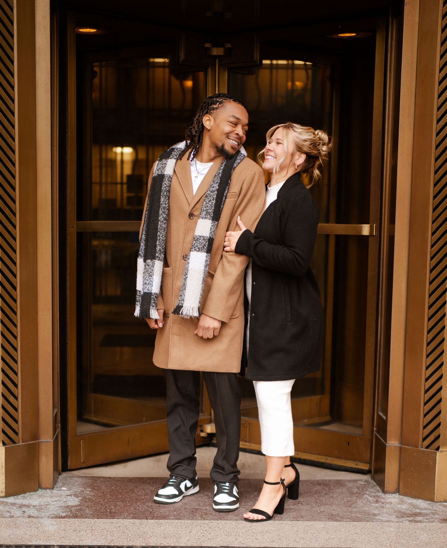 Two people sharing a joyful laugh while standing in a doorway wearing stylish winter coats.