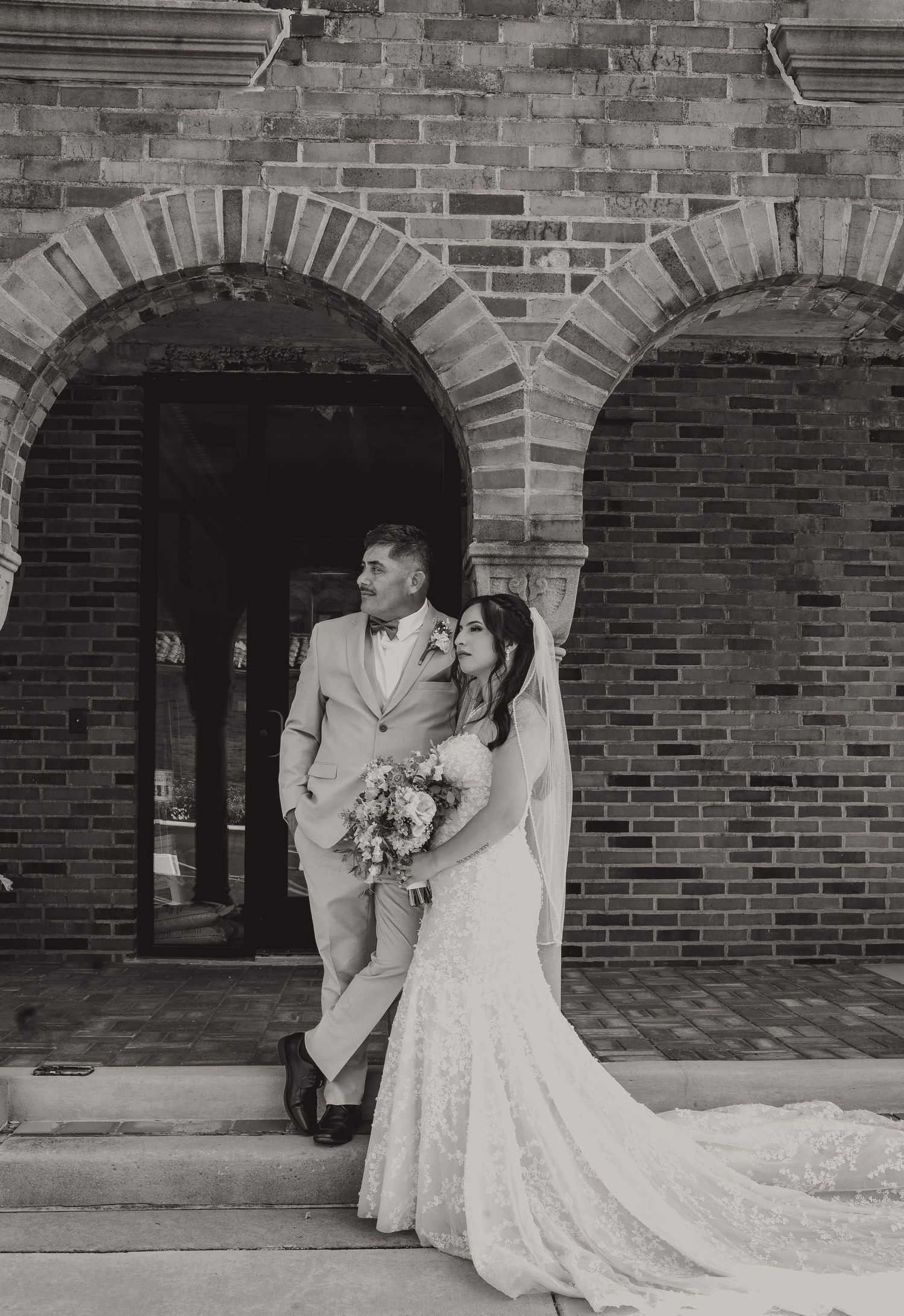 Black and white photo of a bride and groom posing in front of a brick arch doorway on their wedding day.