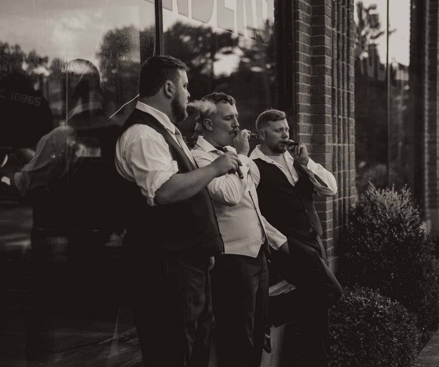 Three men in formal vests stand outside a brick building in a classic black and white street photography style.