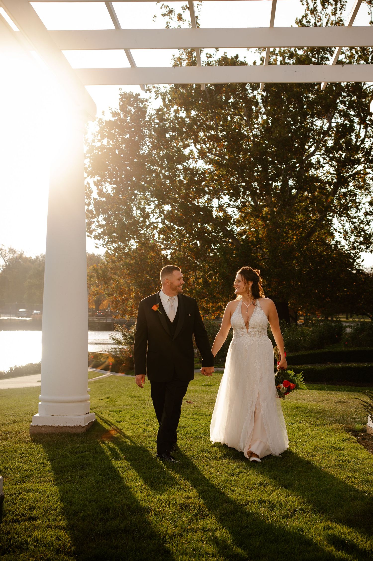 Couple walking hand in hand under pergola at sunset in wedding attire.