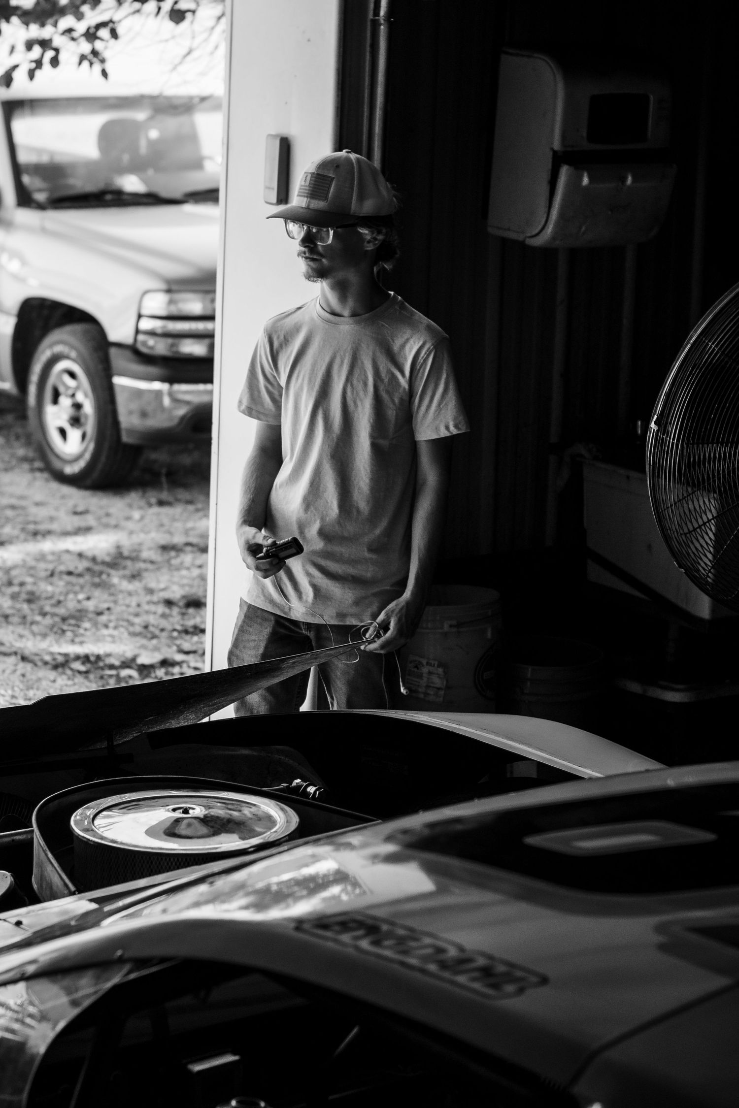 Black and white portrait of someone in a hat standing near a vehicle in a garage setting.
