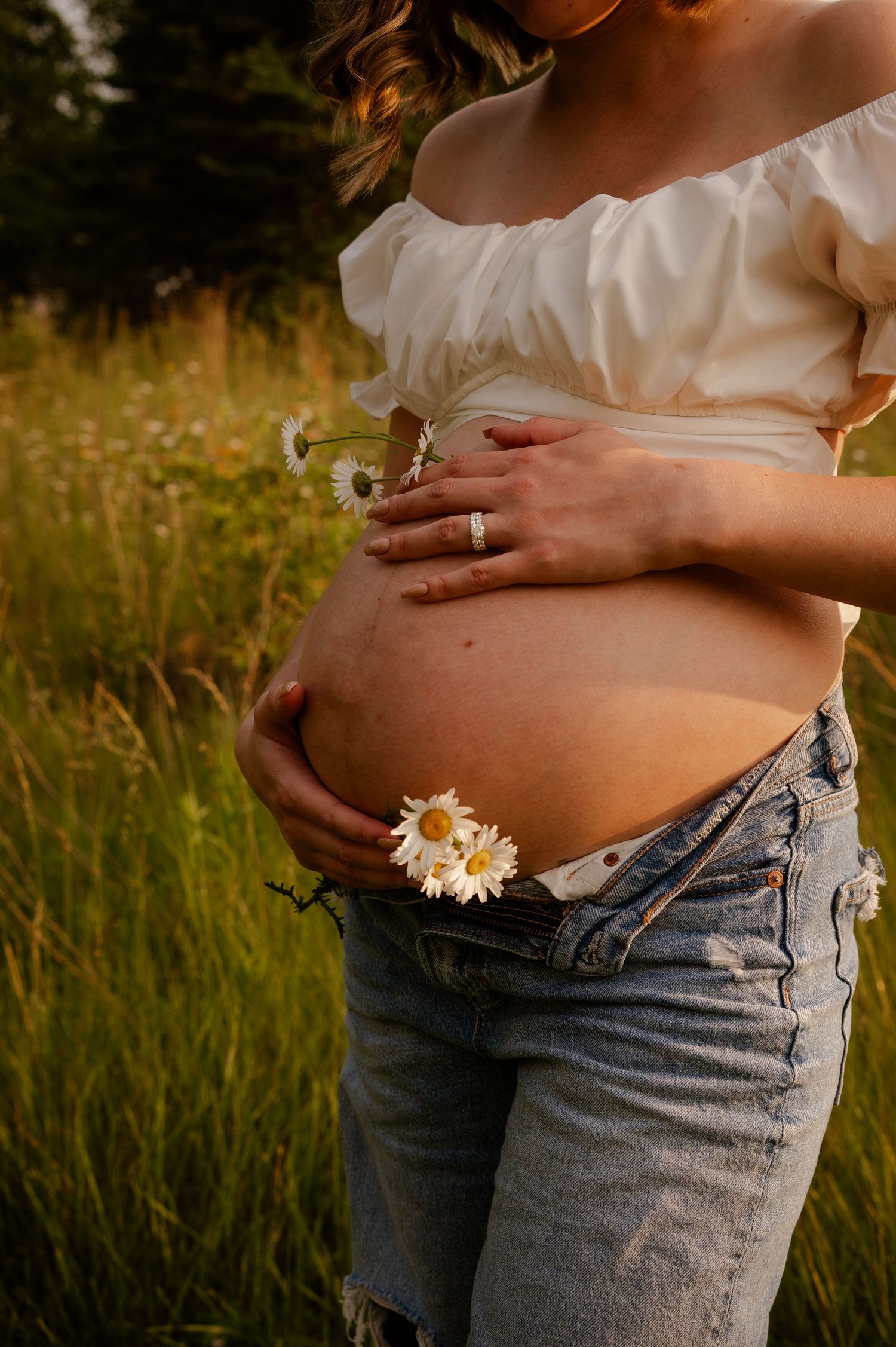 Pregnant woman in white top and jeans holds daisy flowers against her belly in golden sunlit meadow.