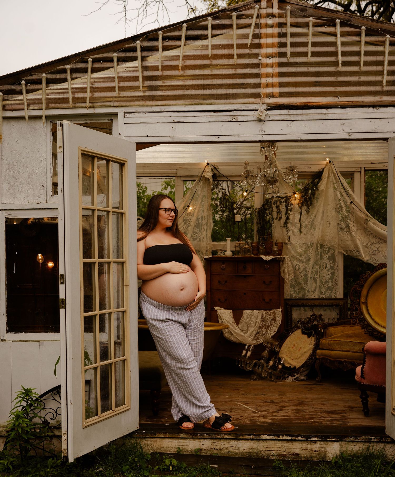 Pregnant woman in black crop top and striped pants stands in rustic boho gazebo doorway at dusk.