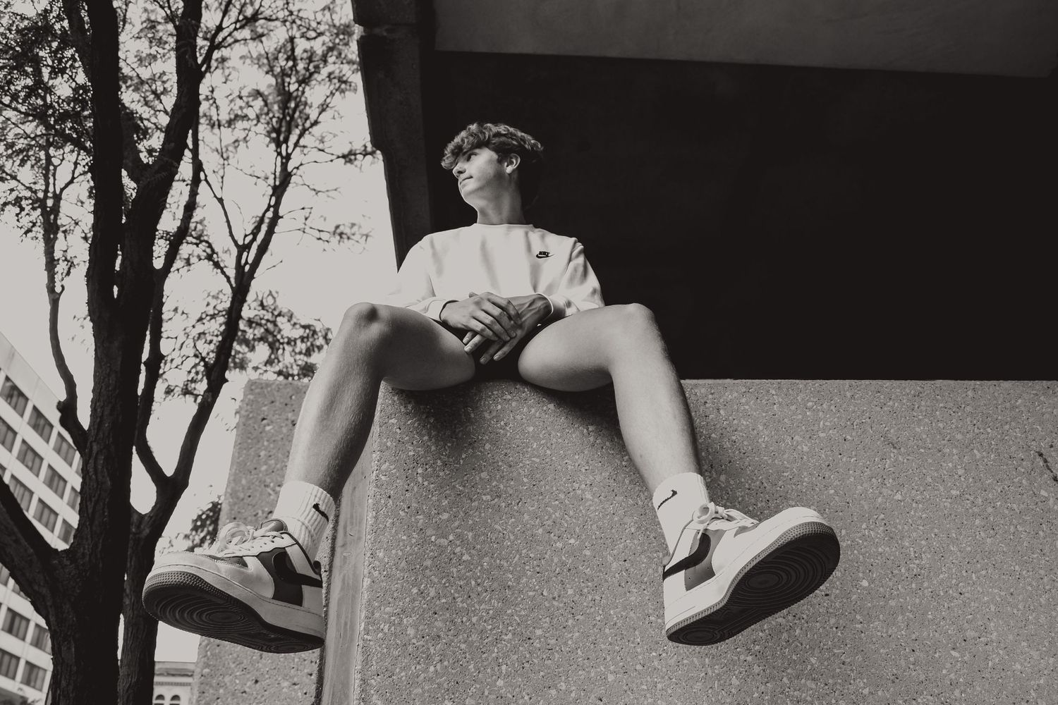 Black and white artistic photo of someone sitting on a ledge against urban architecture.
