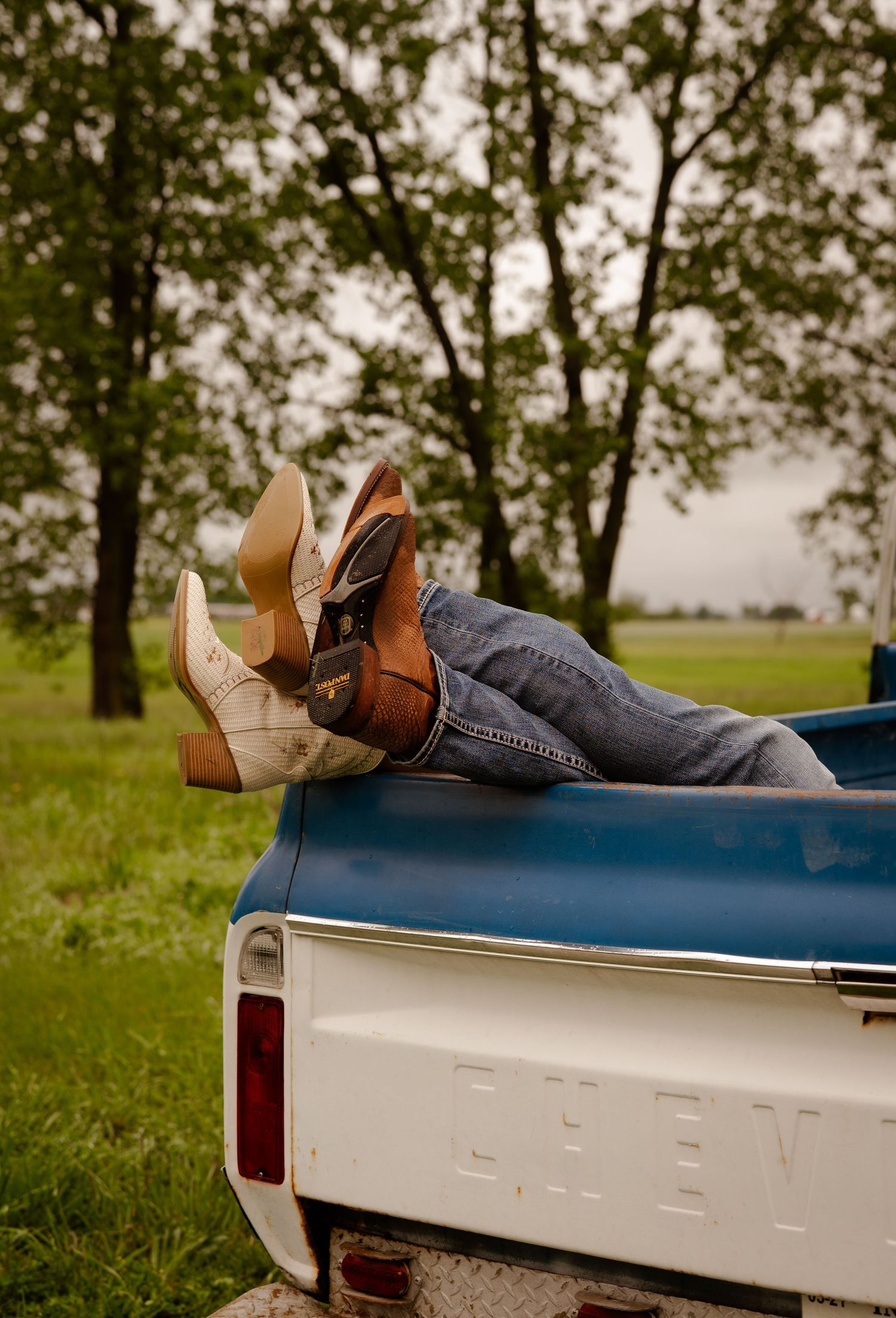 Two people wearing cowboy boots and jeans lying in the bed of a vintage Chevy pickup truck outdoors.